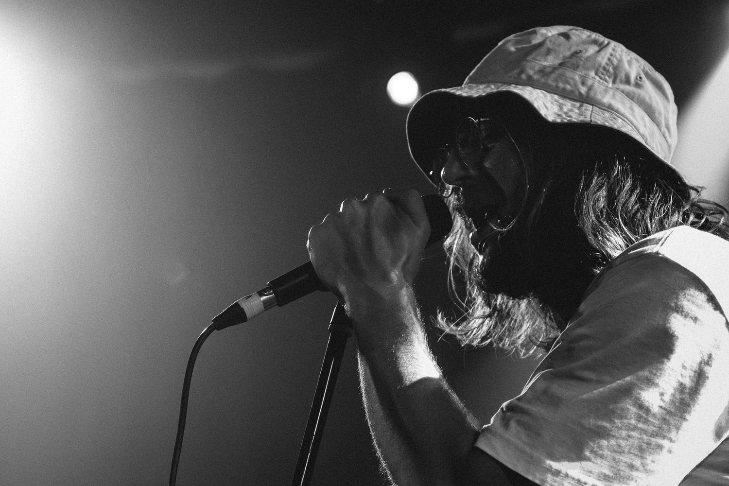 Black and white close-up of lead singer of Winston Surfshirt in bucket hat performing into microphone under stage lights.