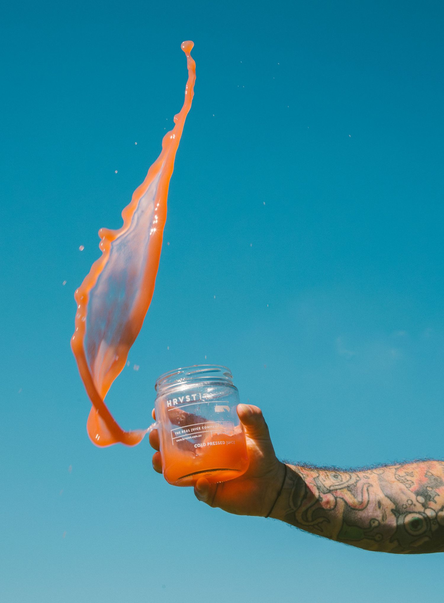 Tattooed hand tossing a HRVST ST jar with a dramatic orange juice splash against a blue sky.