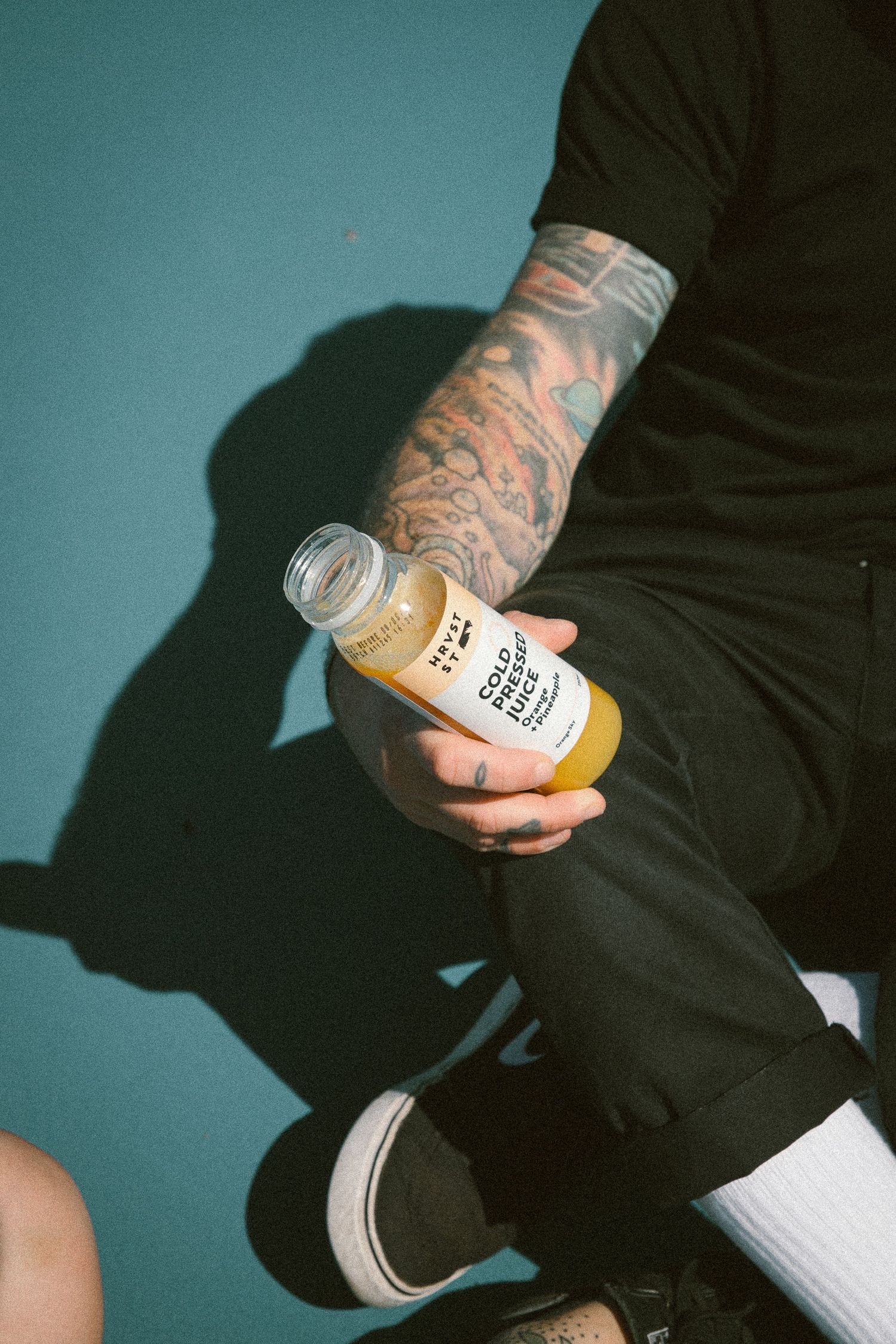 Tattooed man in black outfit holding a HRVST ST Cold Pressed Juice bottle while sitting on a teal surface.