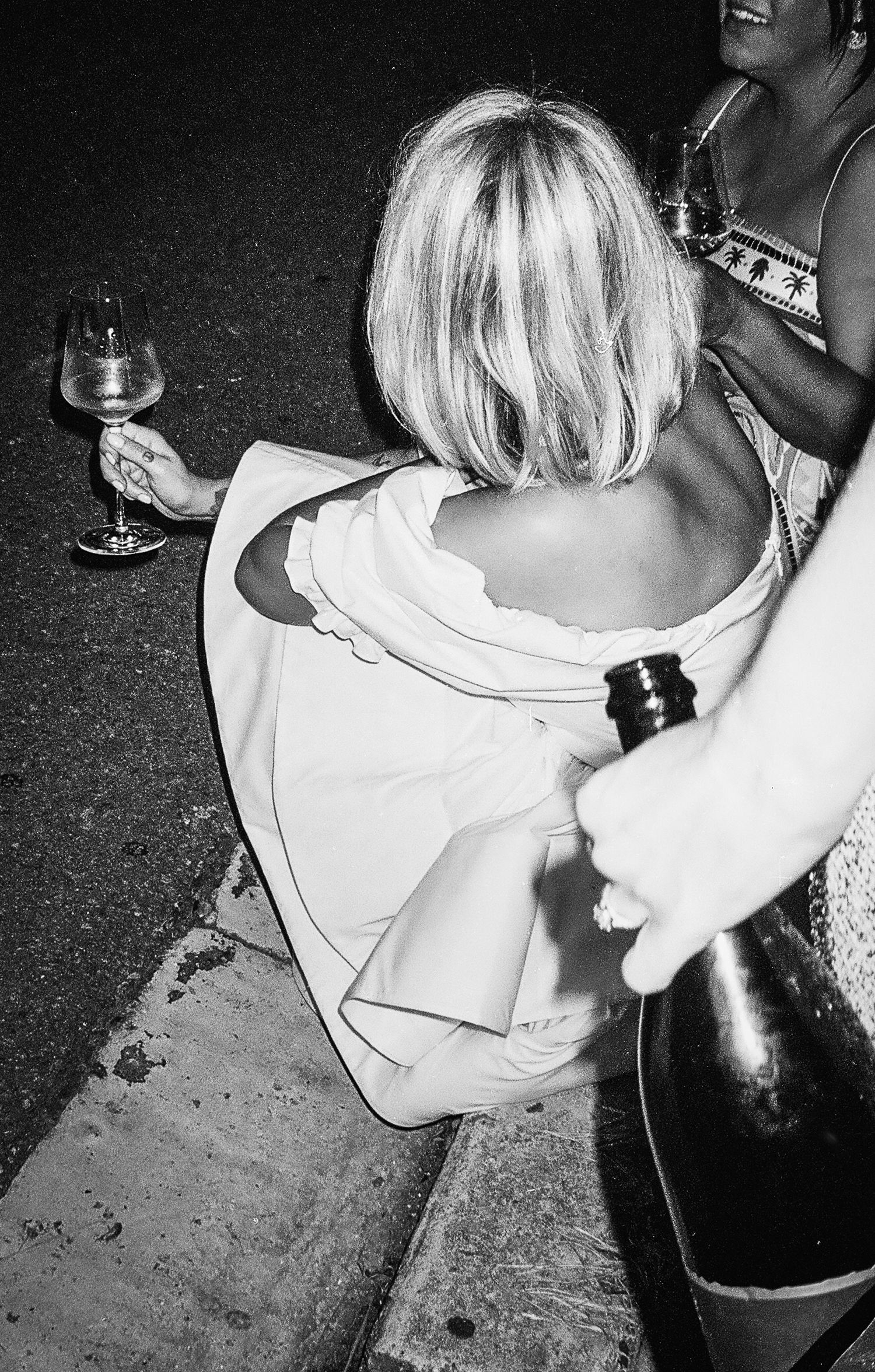 Bride sitting on curb holding wine glass at night, champagne bottle being offered, black and white wedding photo.