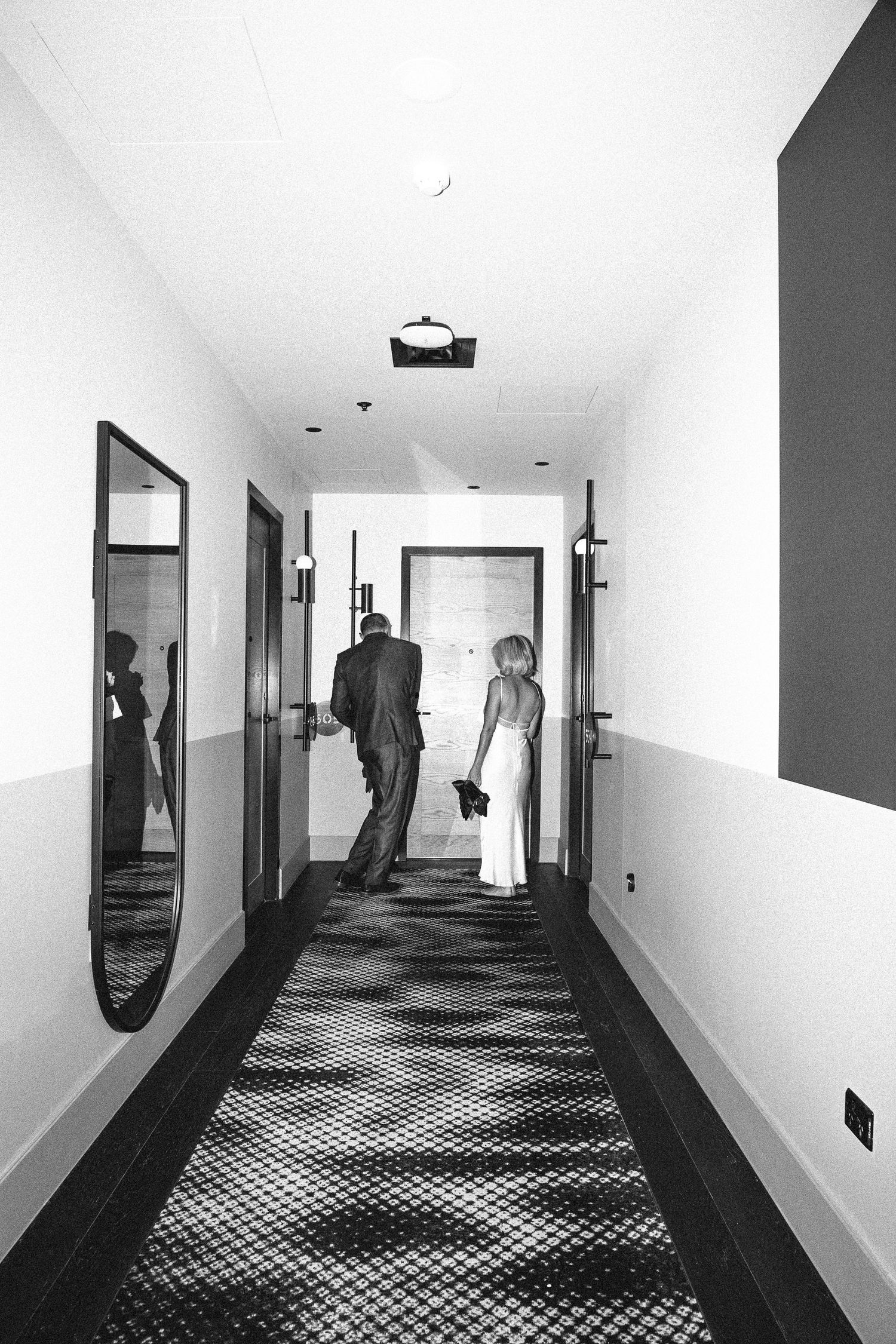 Black and white shot of bride and groom walking down hotel corridor, reflections visible in mirror.