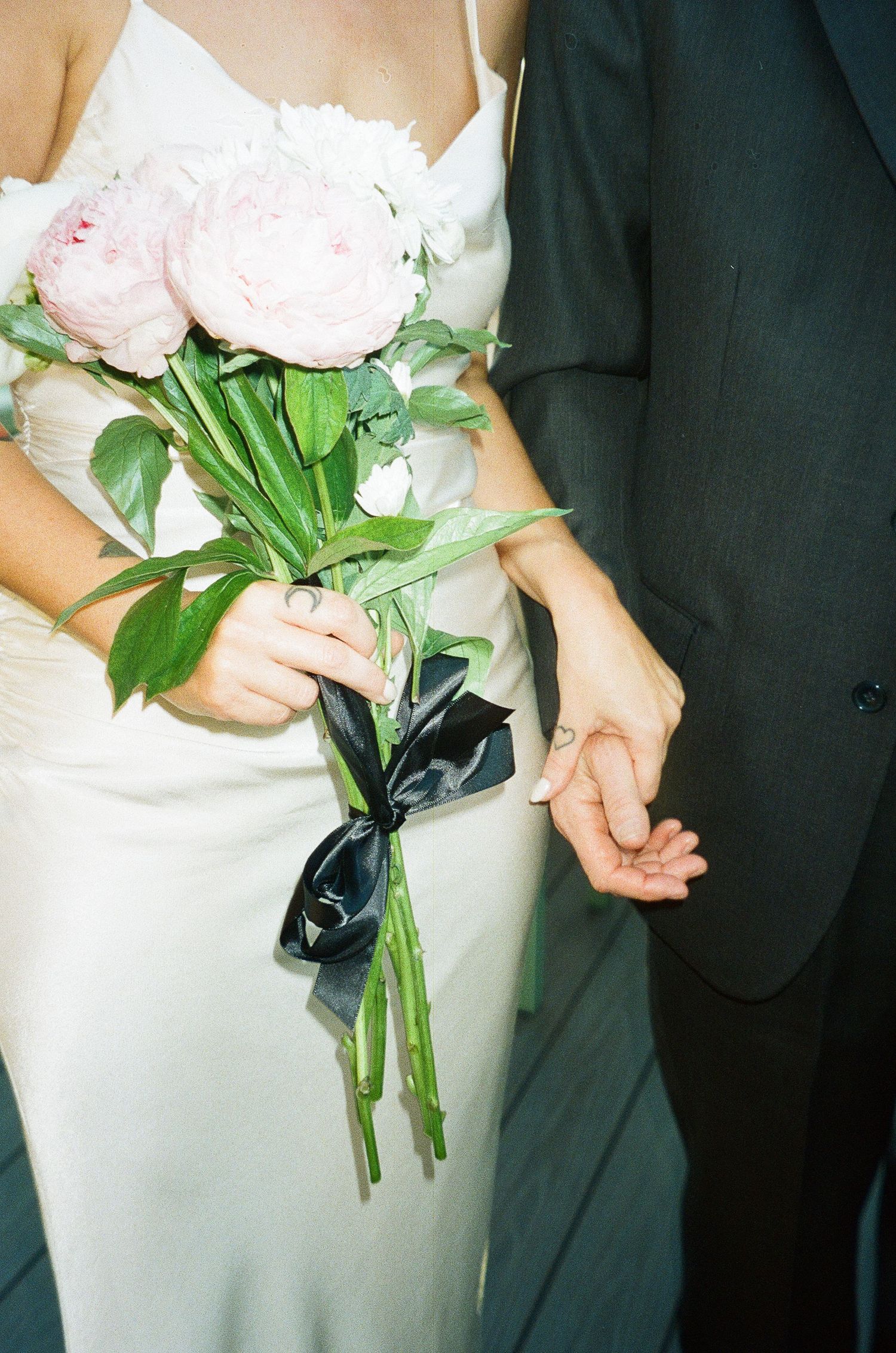 Close-up of bride holding pink peony bouquet tied with black ribbon, groom in dark suit holding hands.