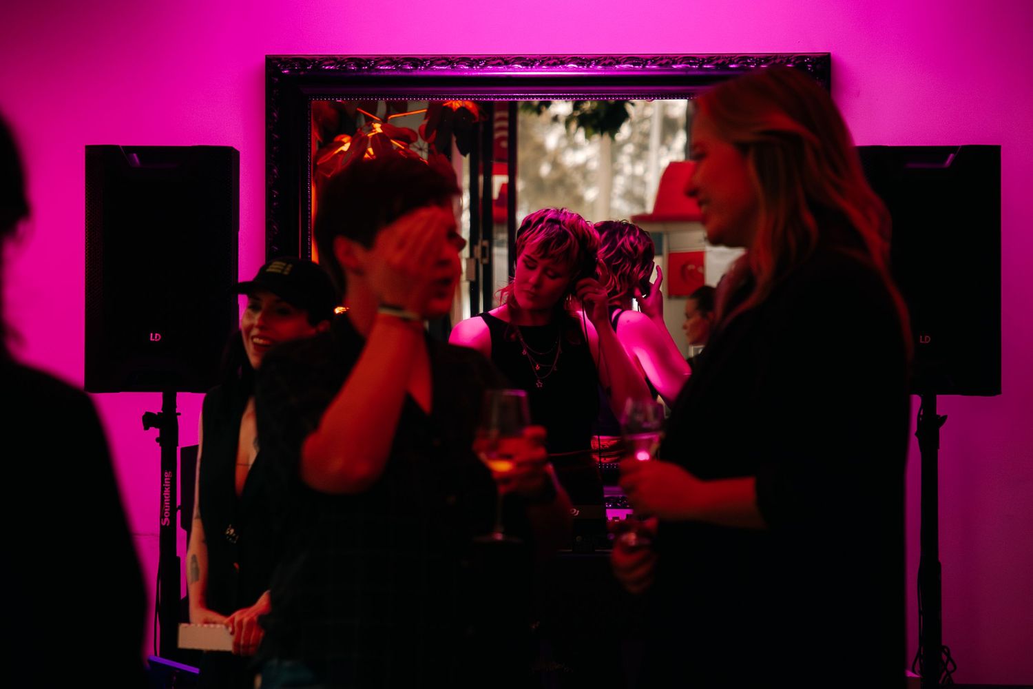 People socializing in pink-lit venue with speakers and mirror at nighttime event.