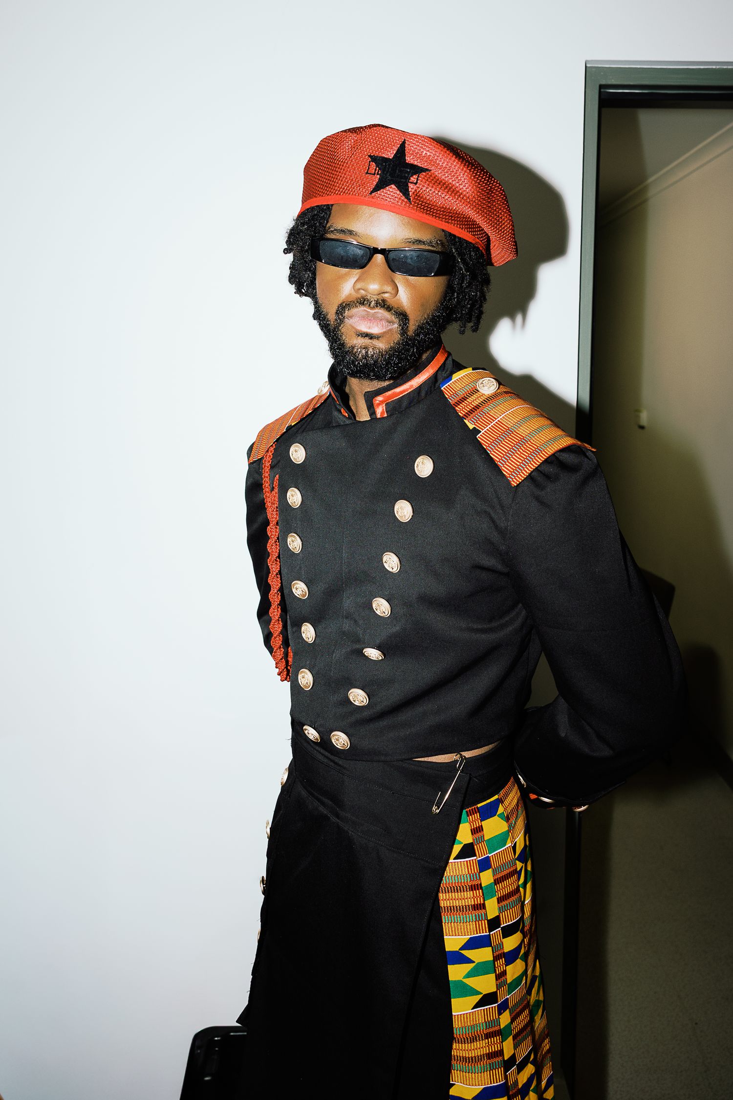 Man in black military jacket, red star beret, sunglasses, and kente cloth skirt posing confidently against white wall.