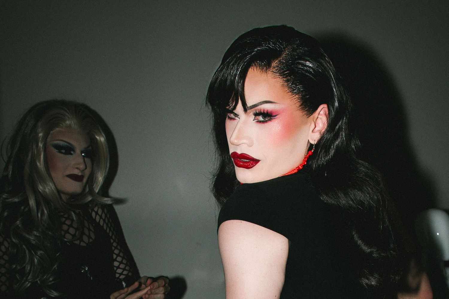 Drag queen with long black hair and bold red lips poses dramatically in dark atmospheric backstage setting The Lass O'Gowrie Hotel.
