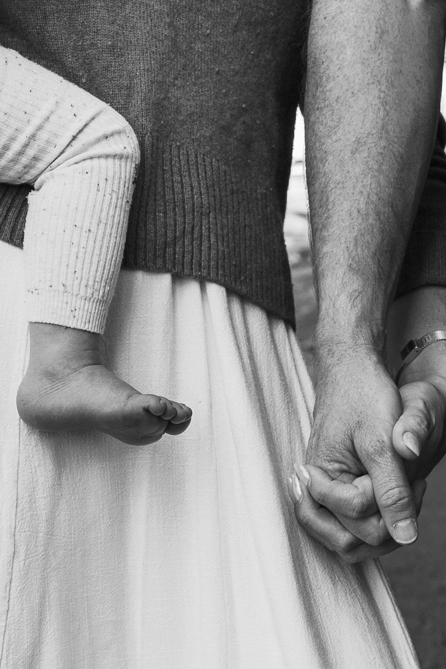 Black and white photo of a couple holding hands with a baby's foot visible, capturing a tender family moment.