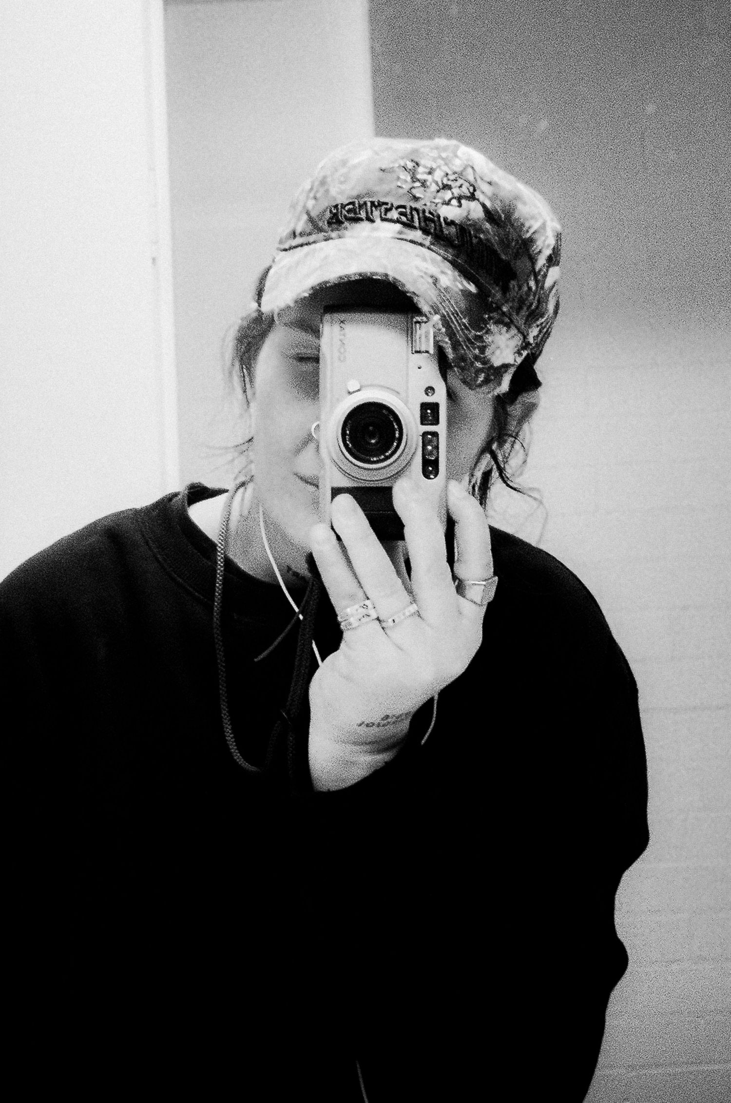 Black and white mirror selfie of person in cap and sweatshirt holding camera covering their face.