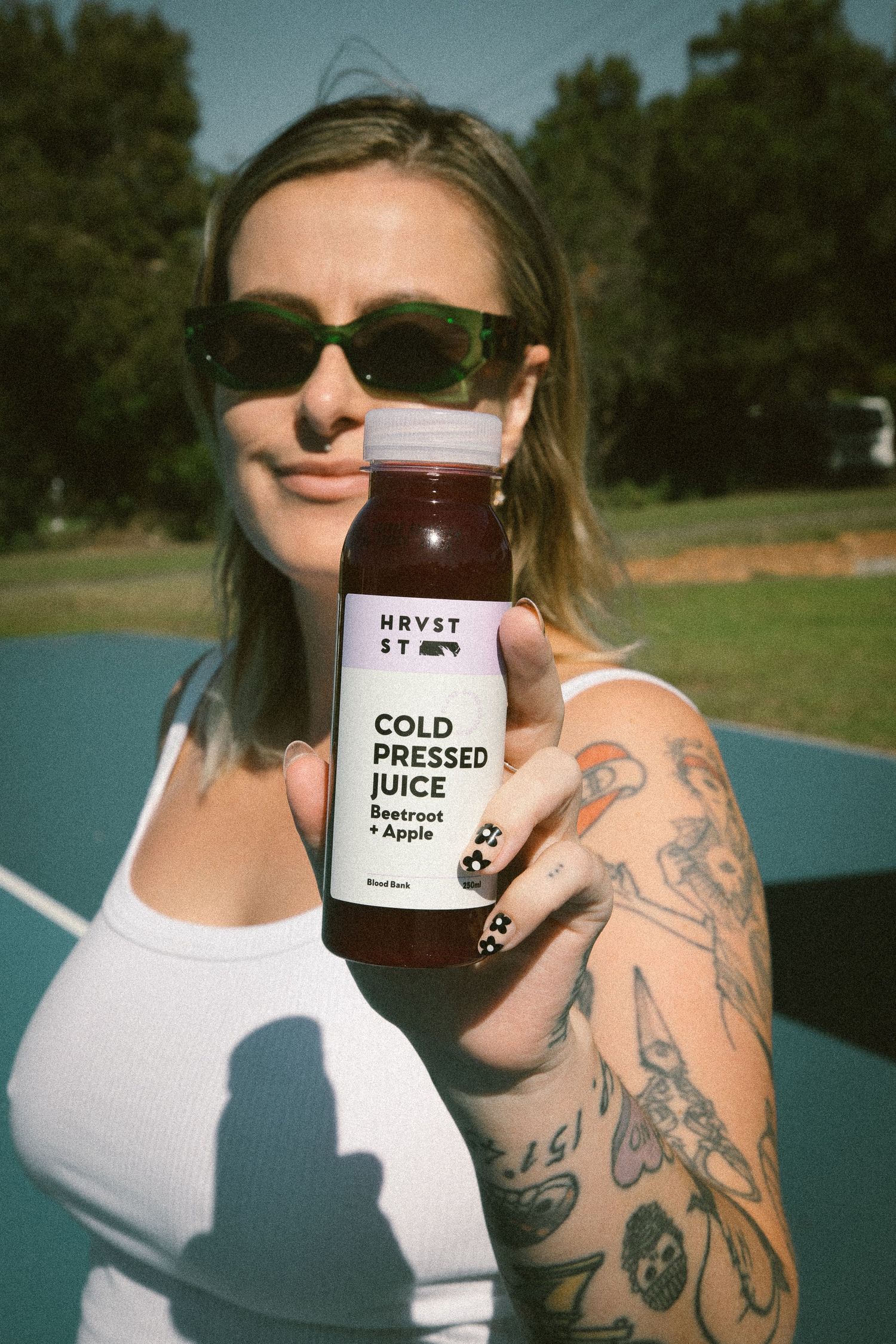 Smiling woman in sunglasses holding up a HRVST ST Cold Pressed Juice Beetroot and Apple bottle outdoors.