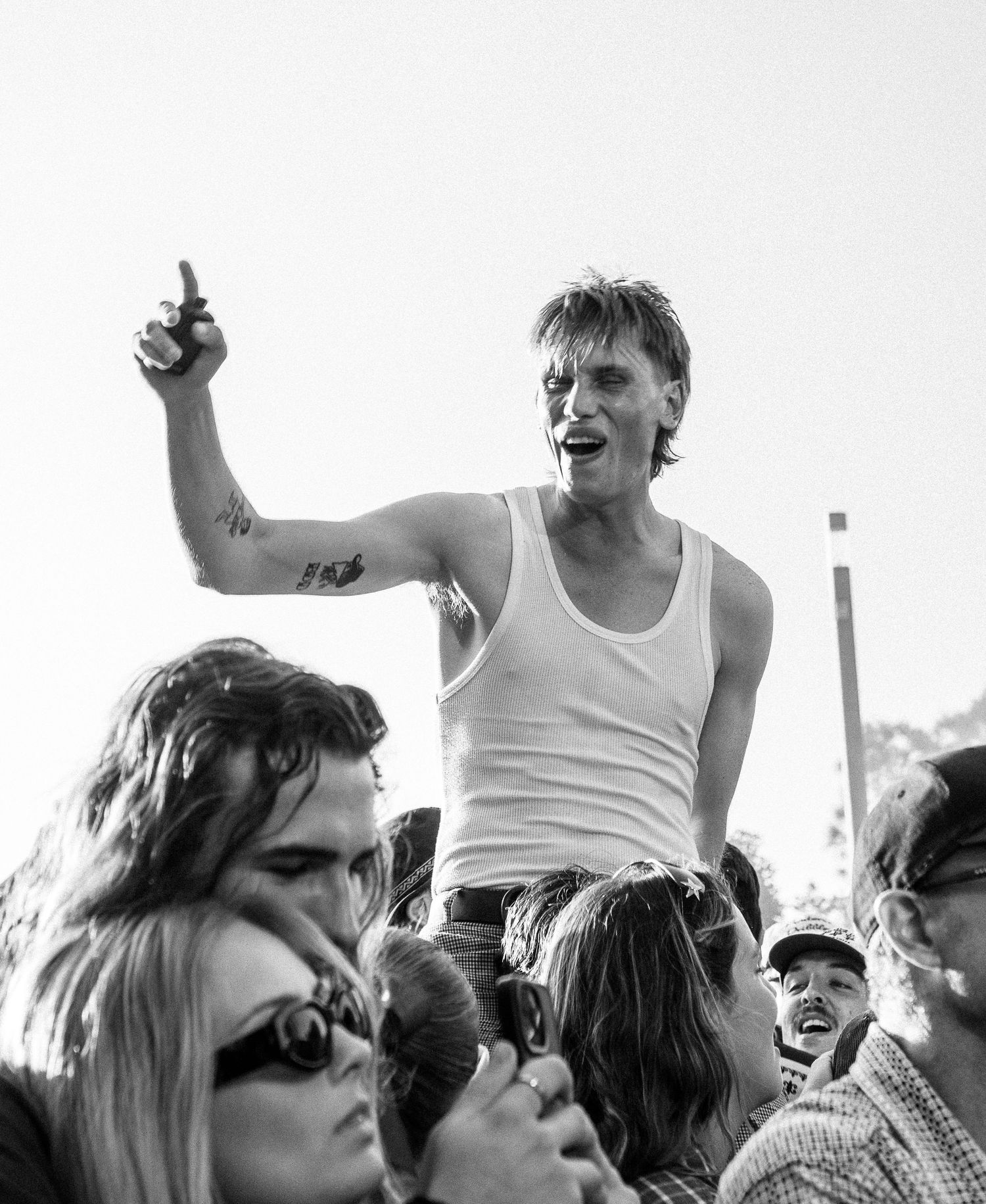 Young man in white tank top crowd surfing at outdoor music Earpfestival, raising finger to sky.