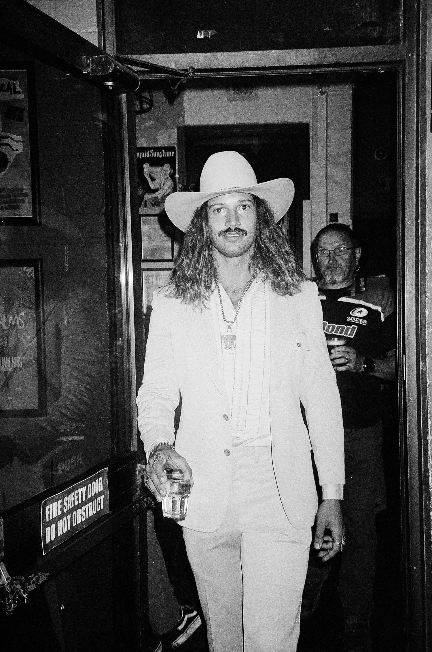 Black and white photo of a long-haired man in a white suit and cowboy hat holding a drink near a fire safety door.