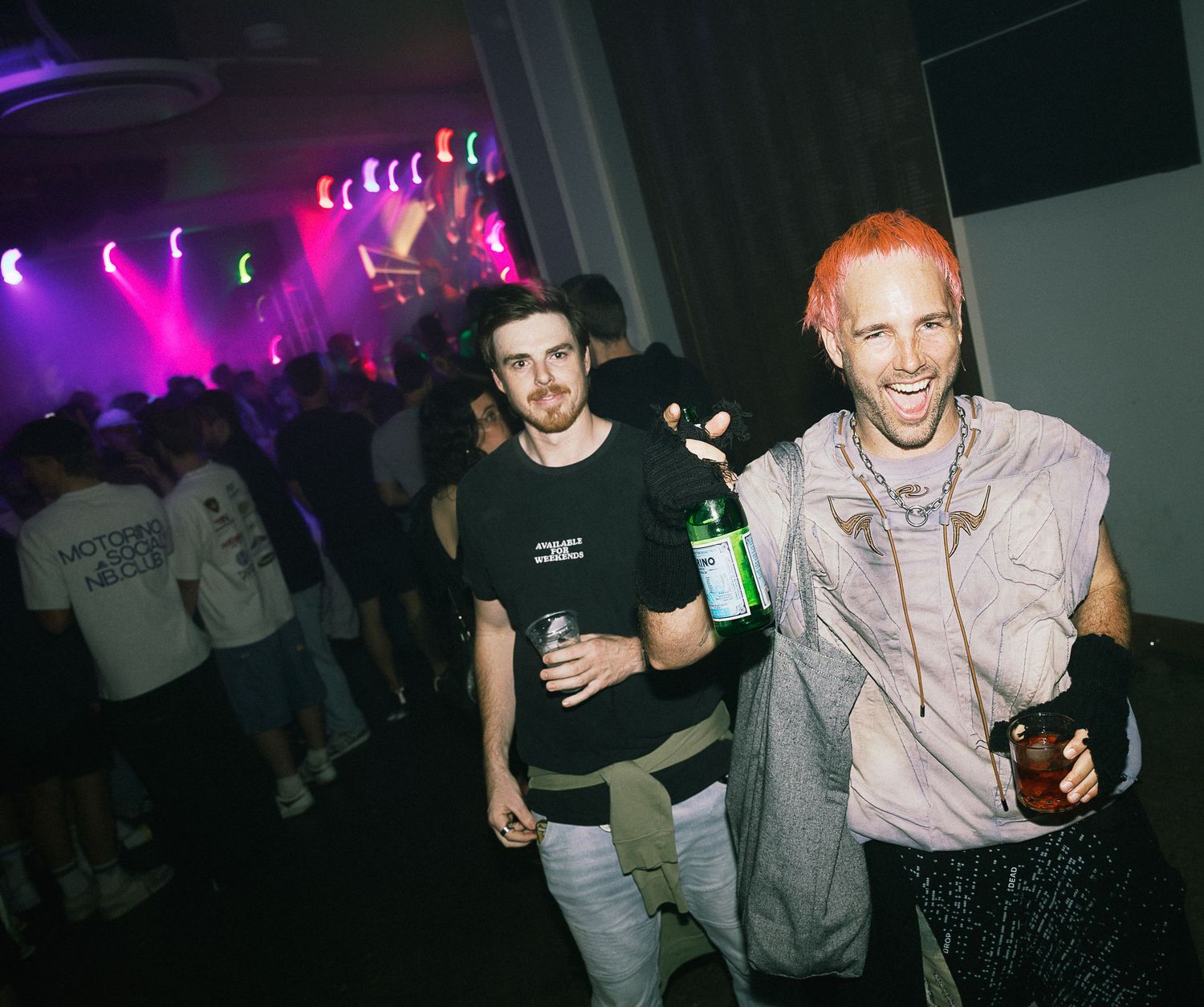 Two men smiling at a vibrant nightclub party, one with orange hair holding drinks under colorful purple stage lights.