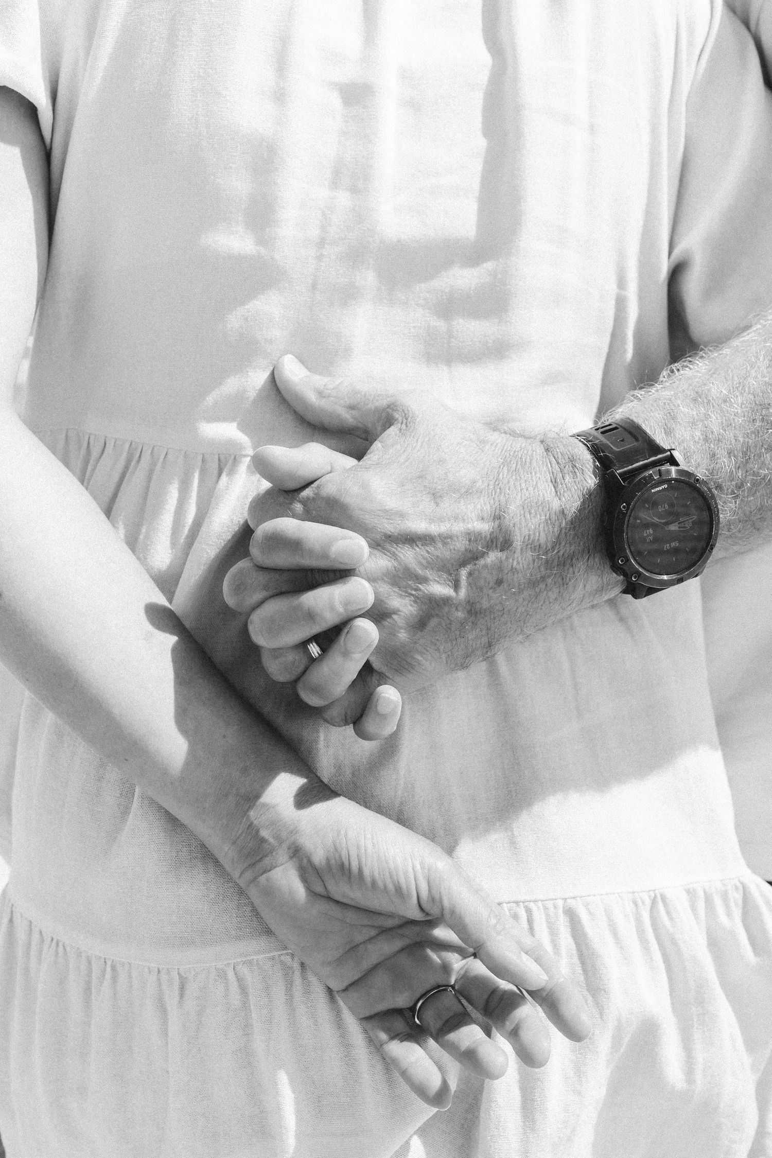 Black and white photo of a couple holding hands, showing wedding rings and a watch, symbolizing love and commitment.