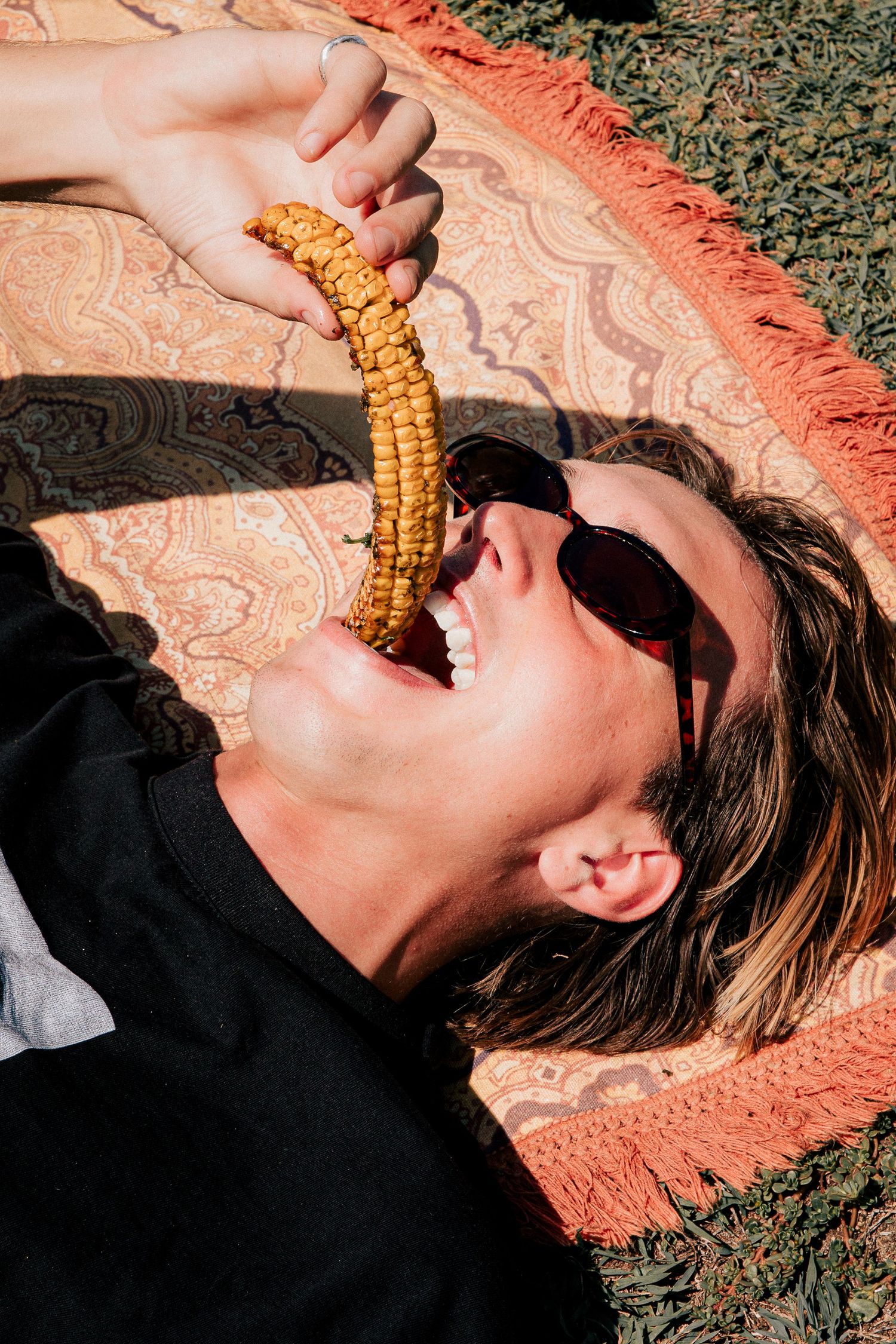 Woman lying on a patterned blanket outdoors, laughing while eating a corn cob and wearing sunglasses at Merewether Beach.
