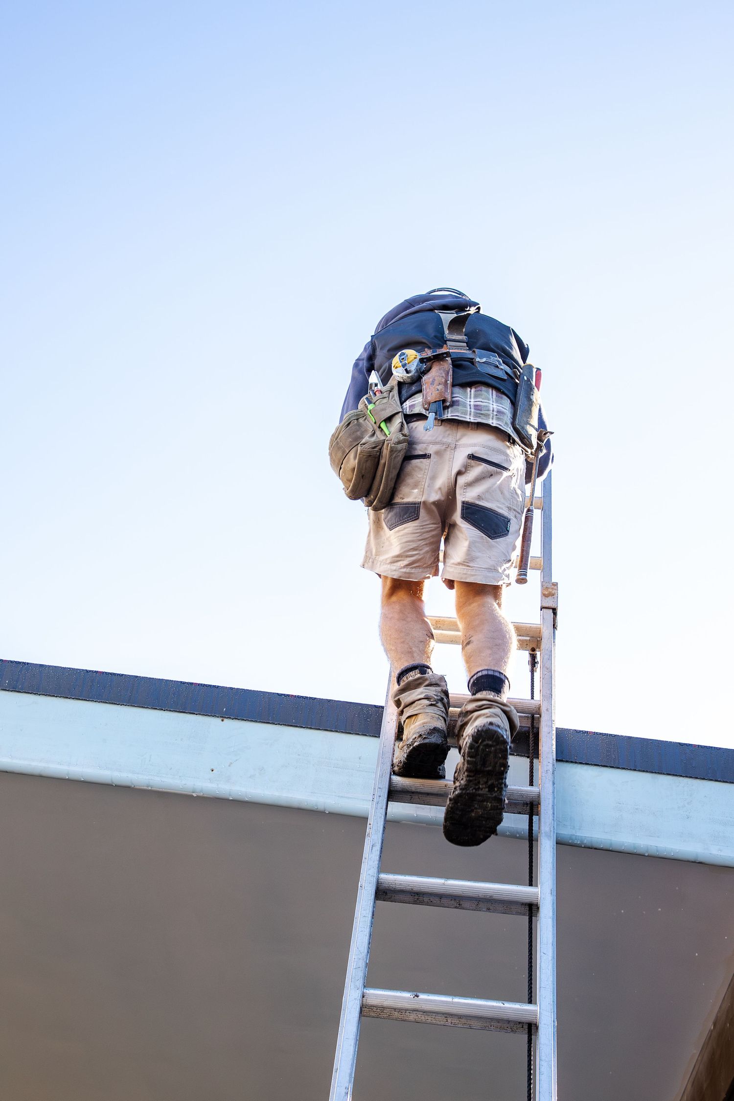 Tradesman climbing a ladder onto a rooftop carrying a tool bag.