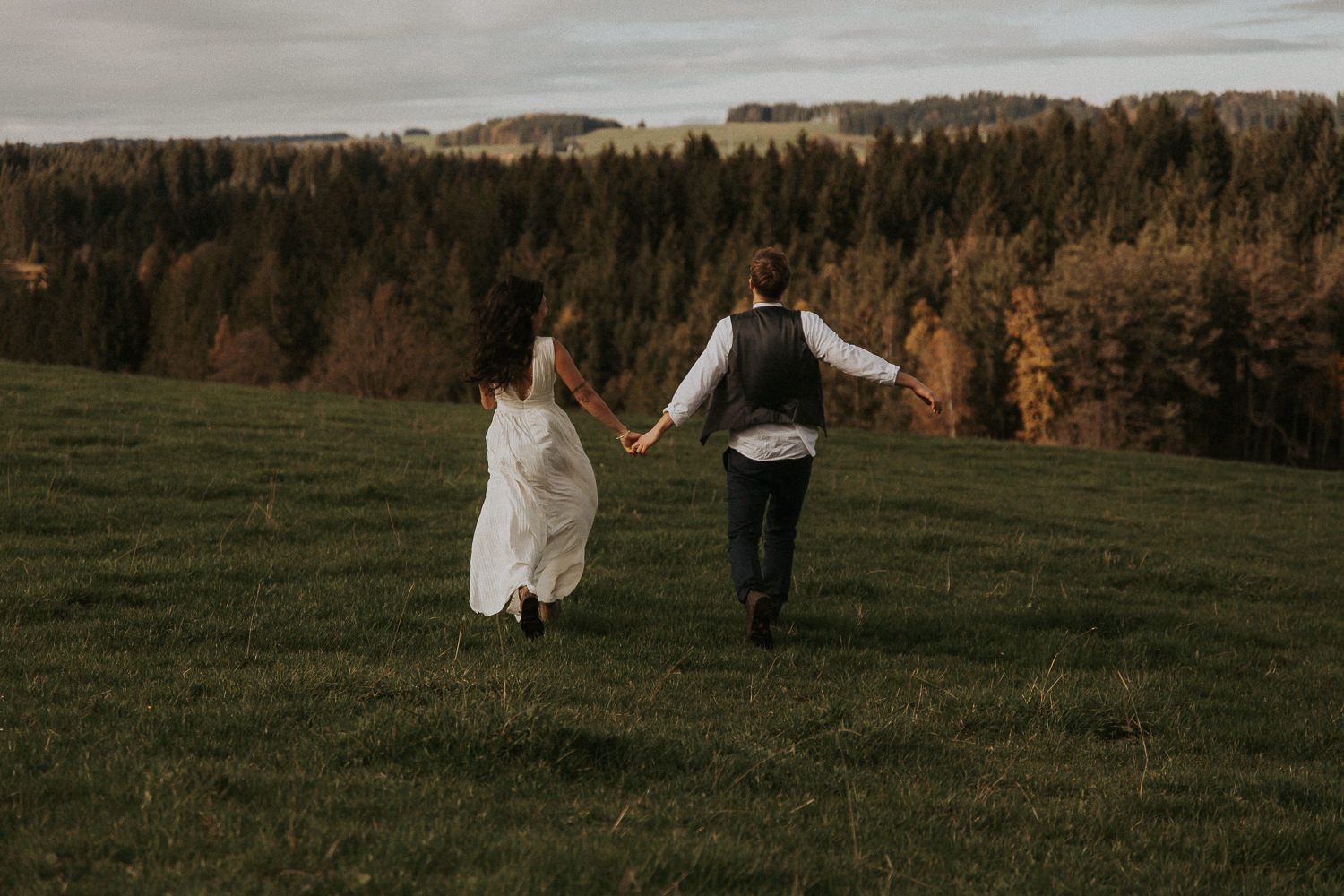 Brautpaar läuft Hand in Hand über eine Herbstwiese mit Waldpanorama