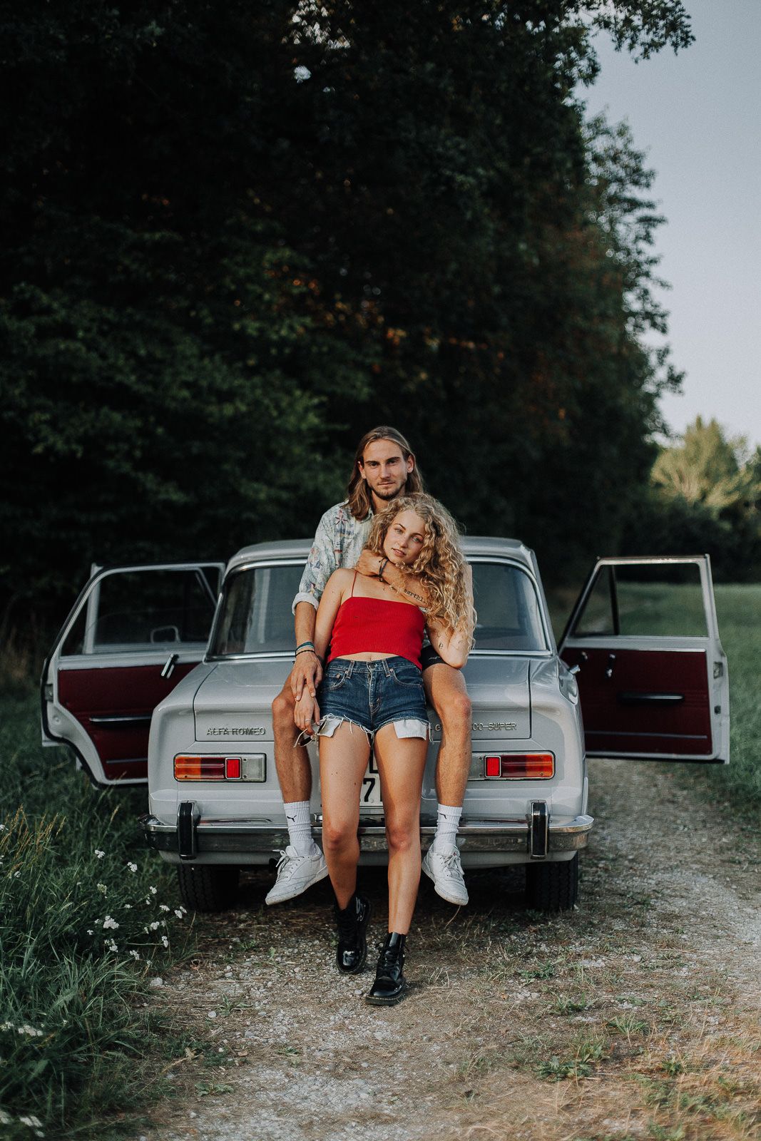 Paar posiert lachend vor einem Oldtimer auf einem Waldweg