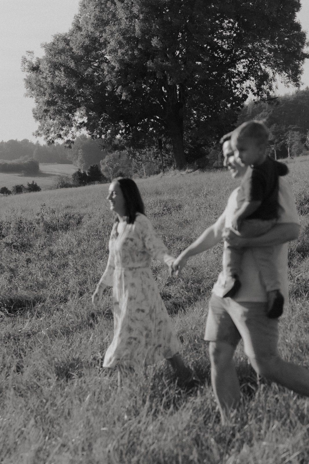 Mutter, Vater und Kind laufen Hand in Hand über eine Sommerwiese – Schwarz-Weiß Familienfotografie