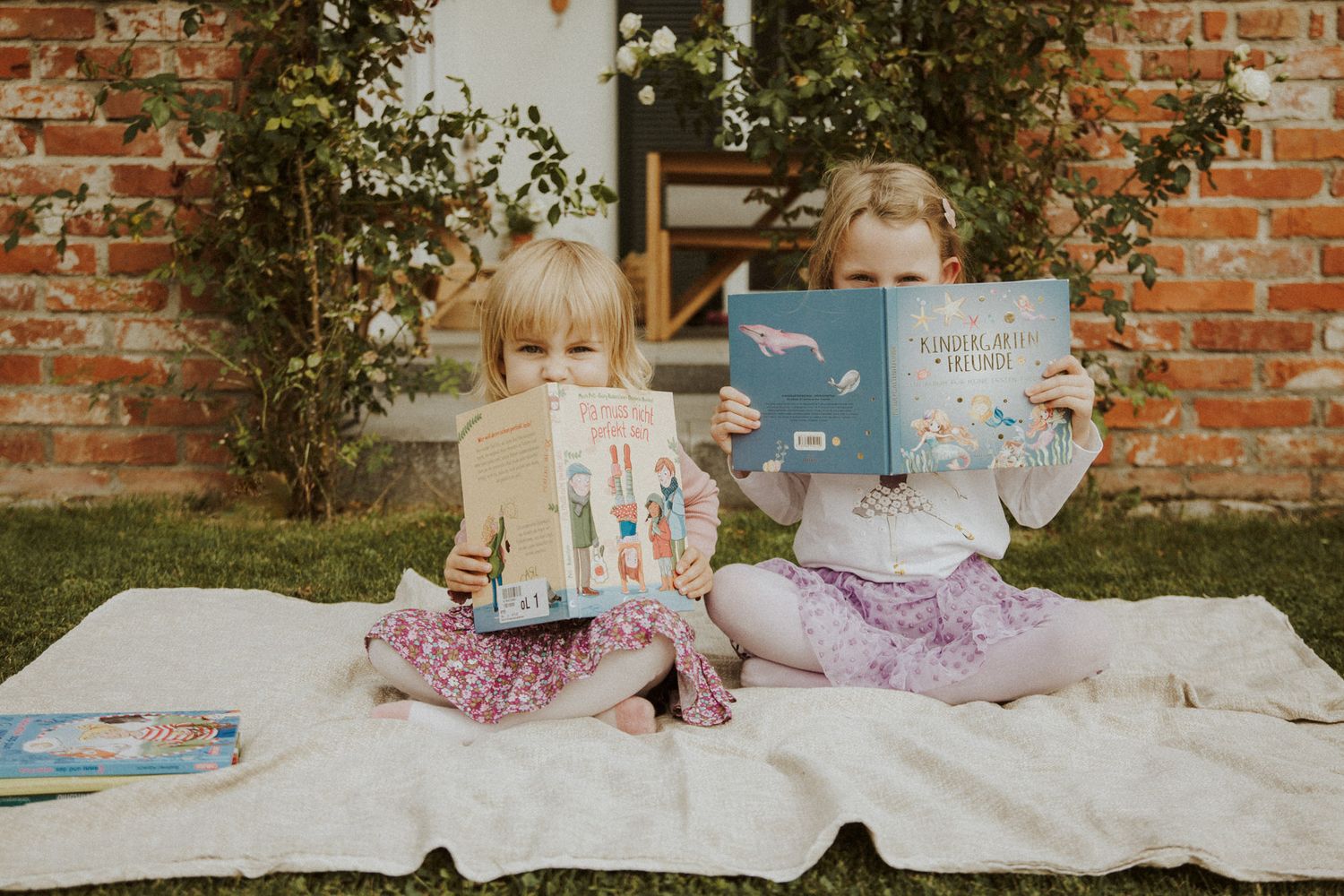 Zwei Schwestern sitzen auf einer Decke im Garten und halten Bücher hoch – Kinderfotografie Ingolstadt