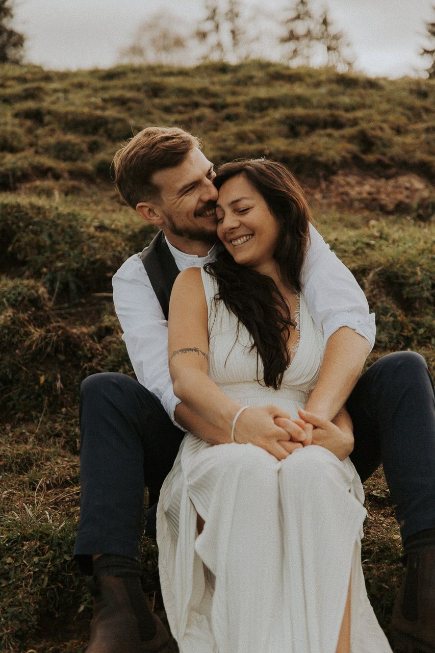 Brautpaar sitzt eng aneinandergekuschelt auf einem Berghang – Hochzeitsfotografie Bayern