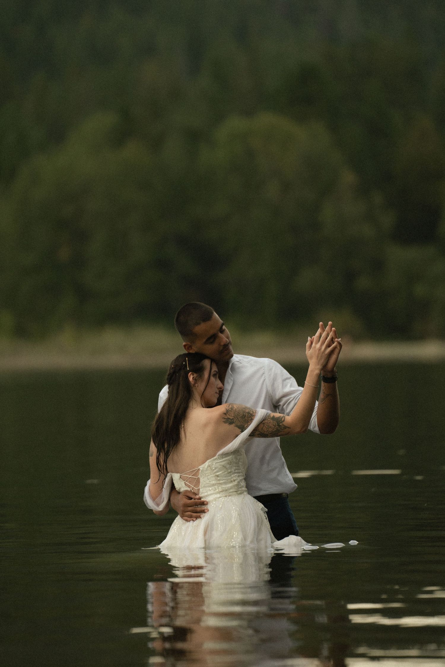 A couple shares a romantic kiss while standing waist-deep in a lake surrounded by lush green trees.