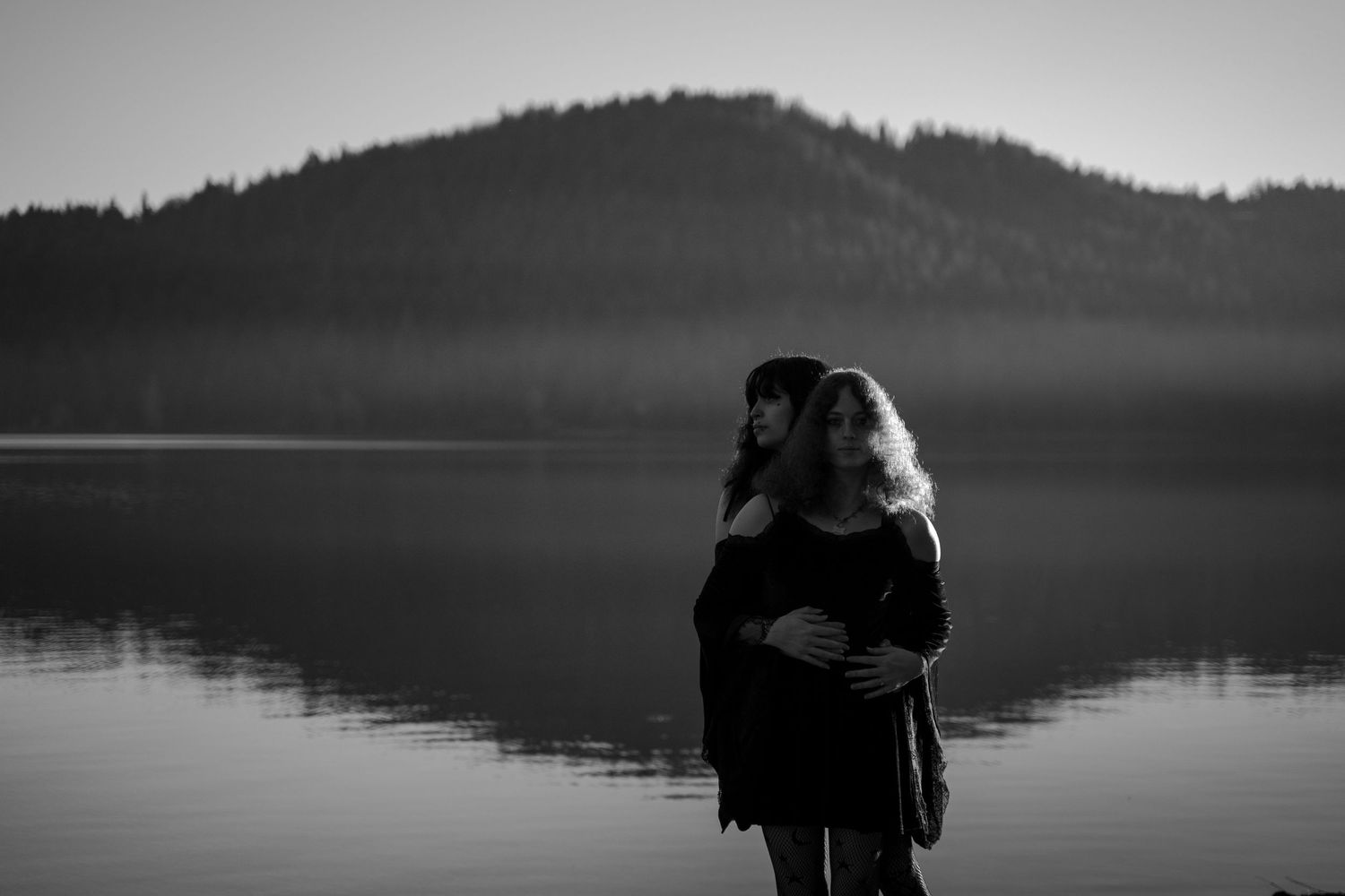 Black and white photo of couple embracing by a misty lake with mountain silhouette backdrop.