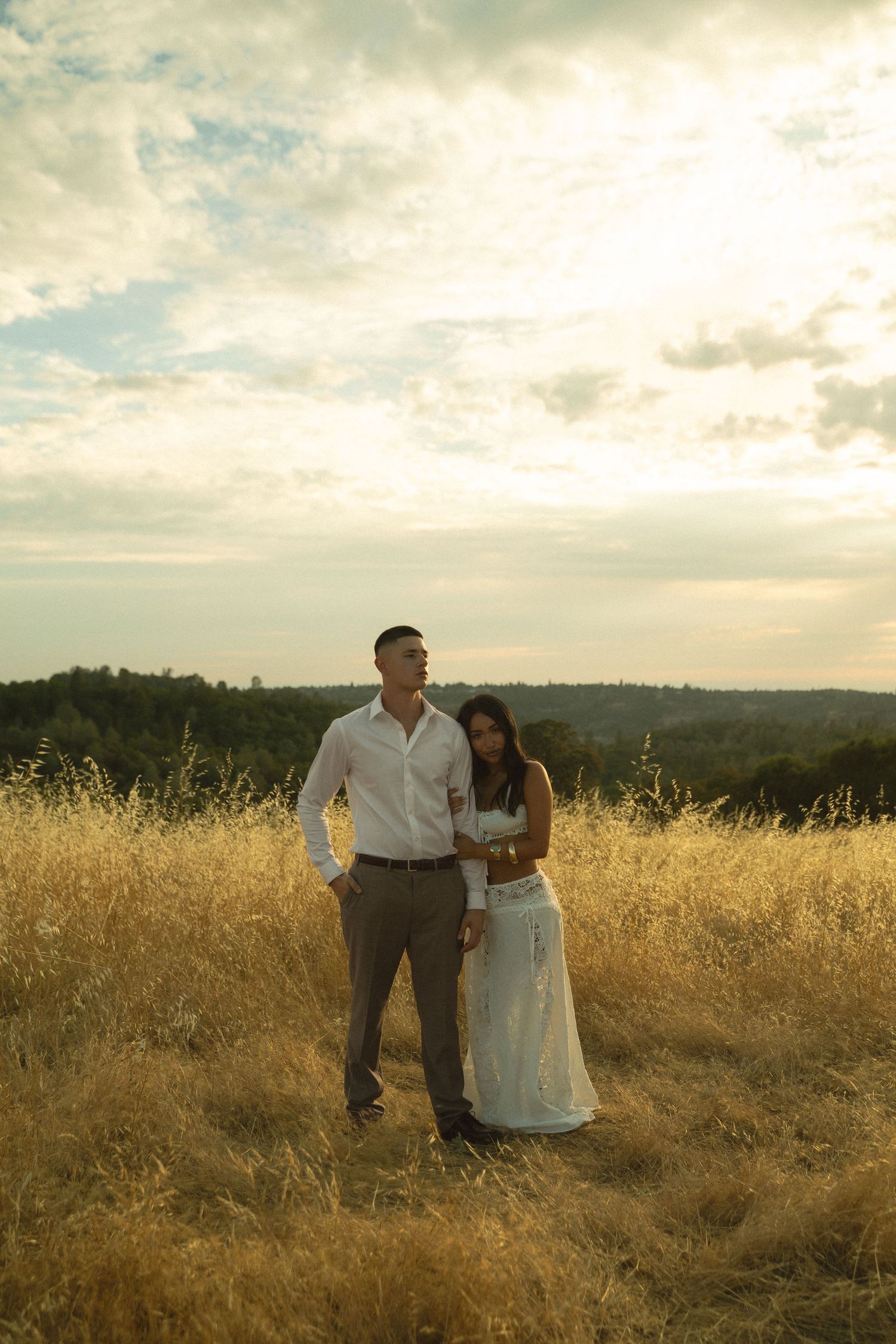 Couple standing together in golden wheat field during glowing sunset.