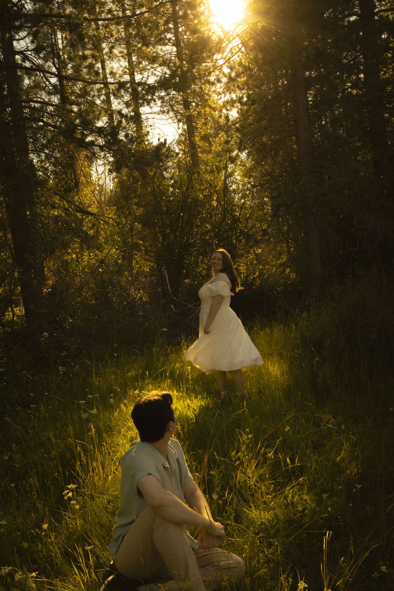 Man sitting in grass watching woman in white dress twirl in sunlit forest.