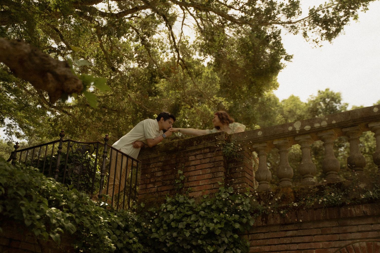 Romantic couple reaching for each other over a brick garden wall surrounded by lush greenery.