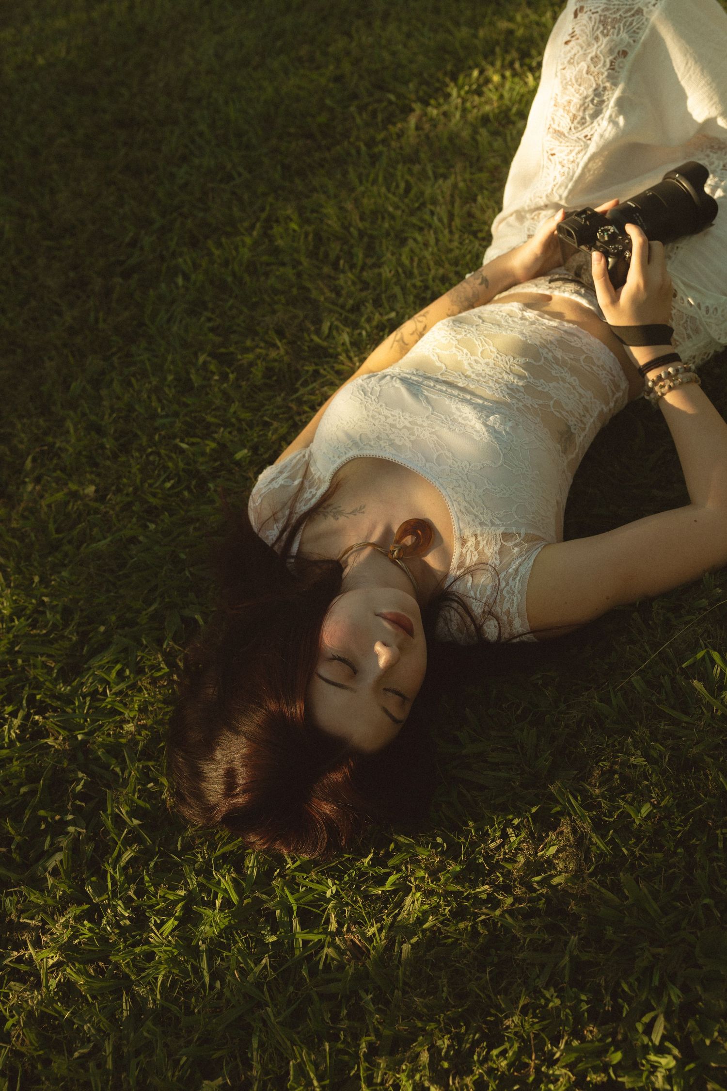 Woman in white lace dress lying on green grass holding camera in warm golden light.