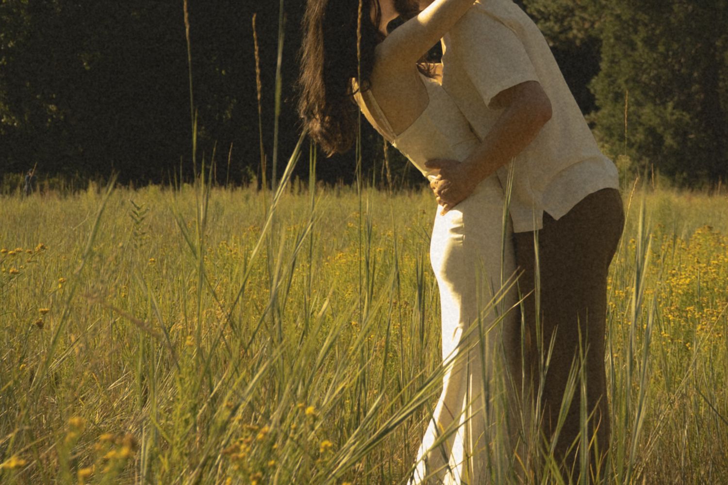 Couple sharing a tender embrace in a sunlit golden meadow surrounded by tall grass and wildflowers.
