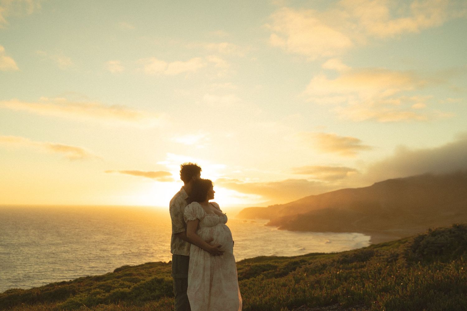 A couple embraces silhouetted against a stunning golden sunset over coastal cliffs and the ocean.