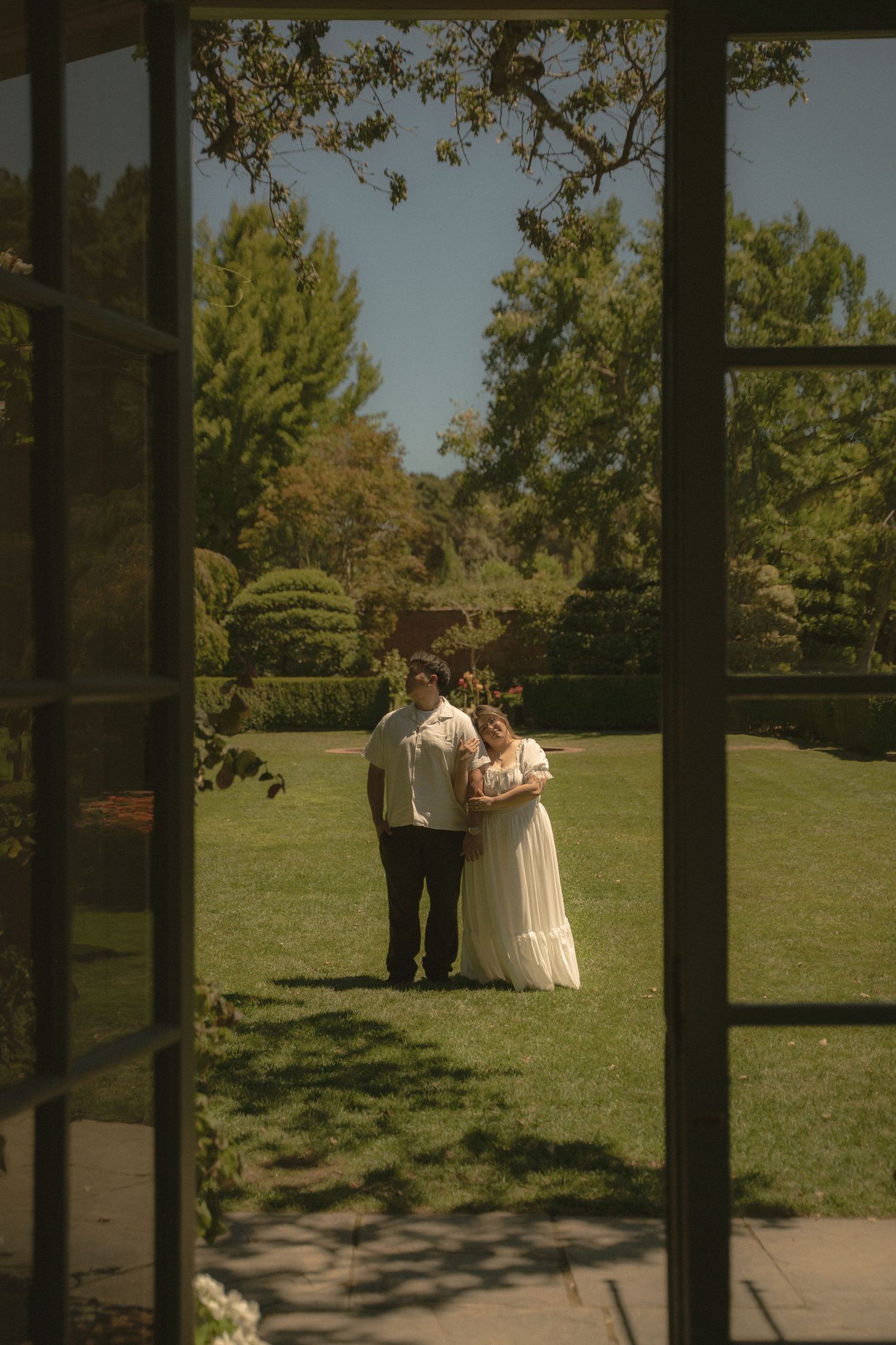 A bride and groom stand together in a lush green garden, viewed through elegant glass French doors.