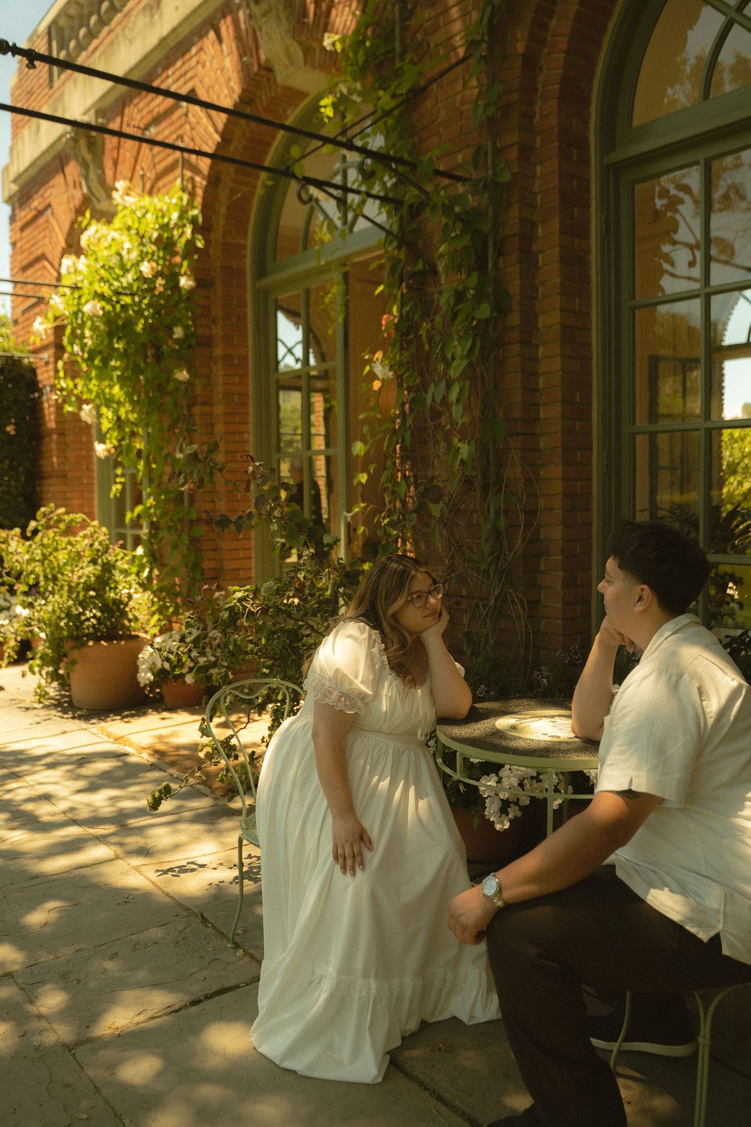 Couple in white attire sitting at outdoor table by ivy-covered brick building in golden sunlight.