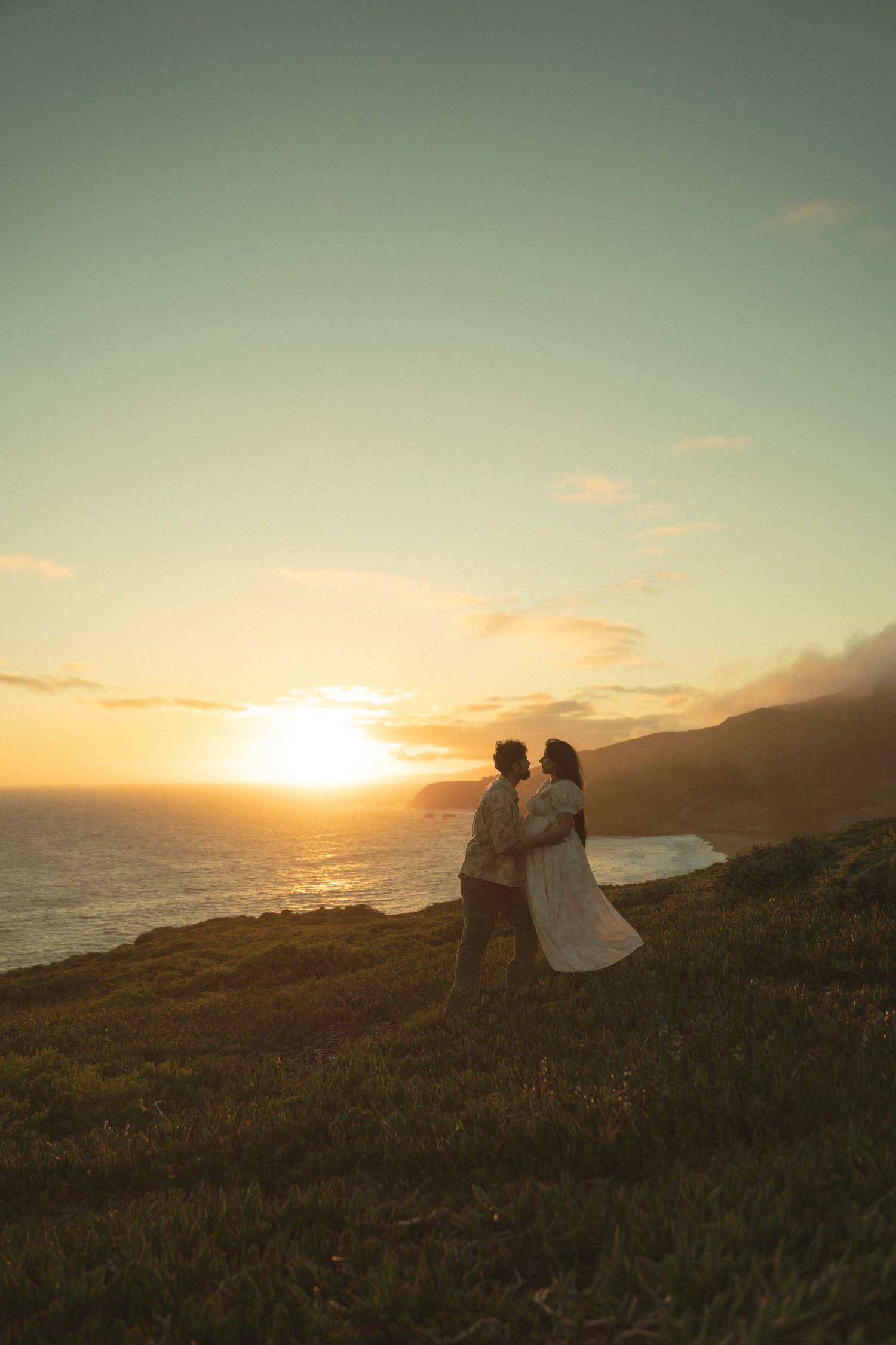 Romantic couple embracing at sunset on a coastal hillside with golden ocean views.