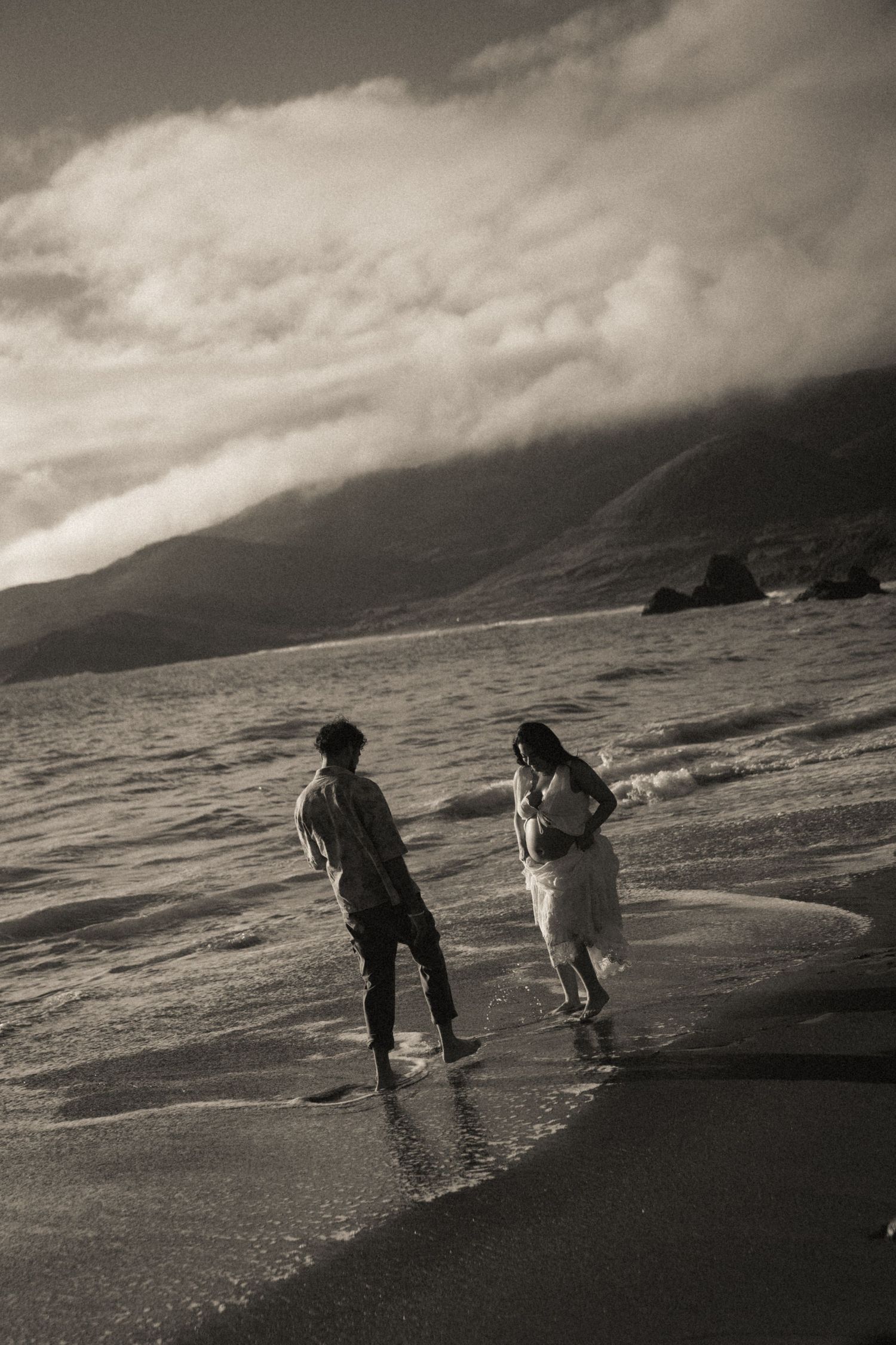 Couple walking along a misty beach shoreline in black and white, with mountains and dramatic clouds in the background.