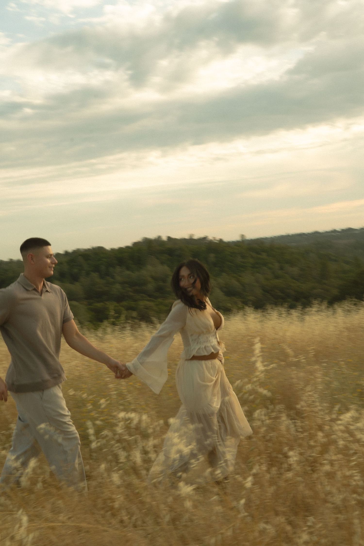 Couple holding hands walking through golden field under cloudy sky.