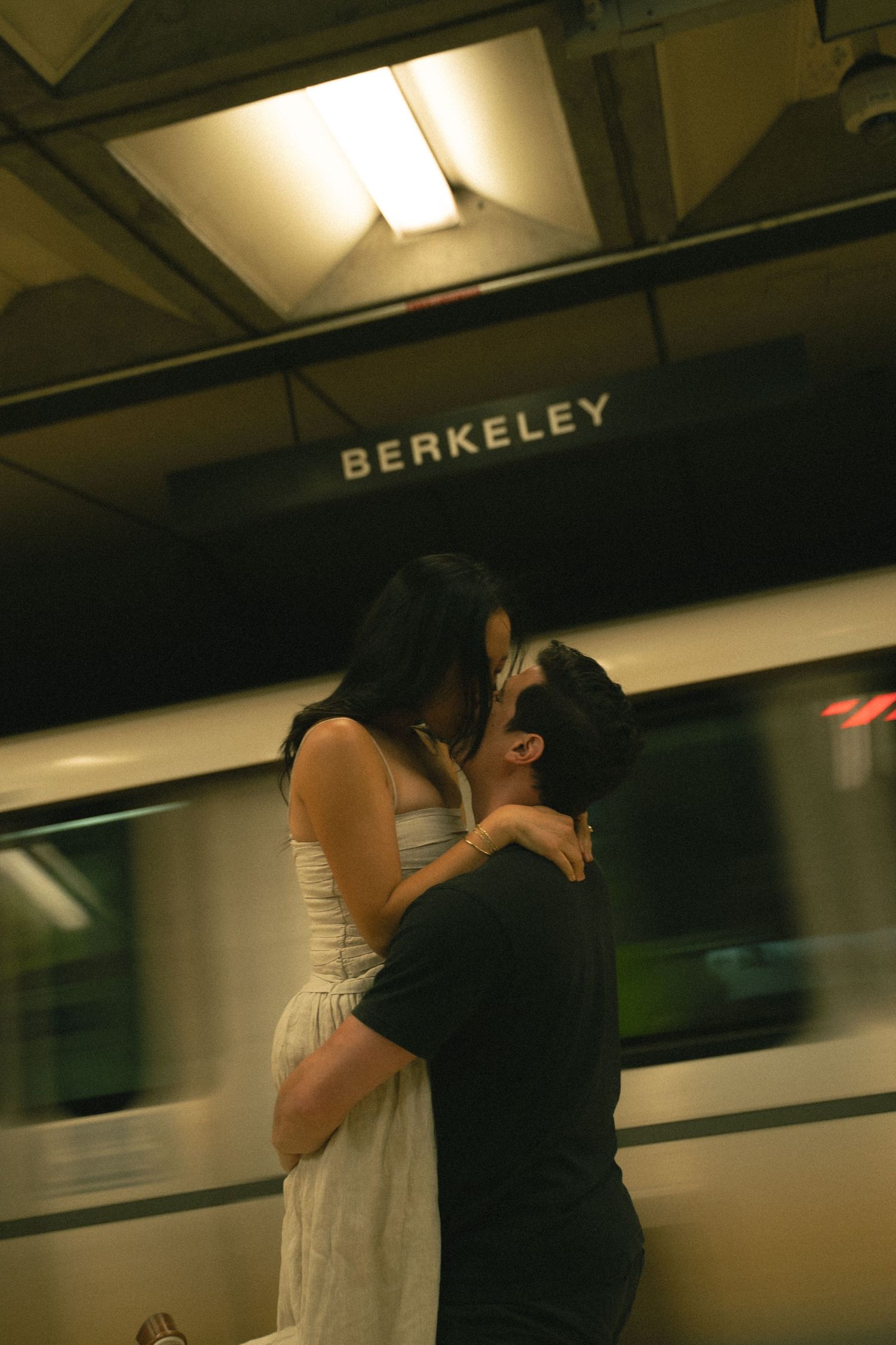 Couple sharing a passionate kiss at Berkeley subway station with speeding train in background.
