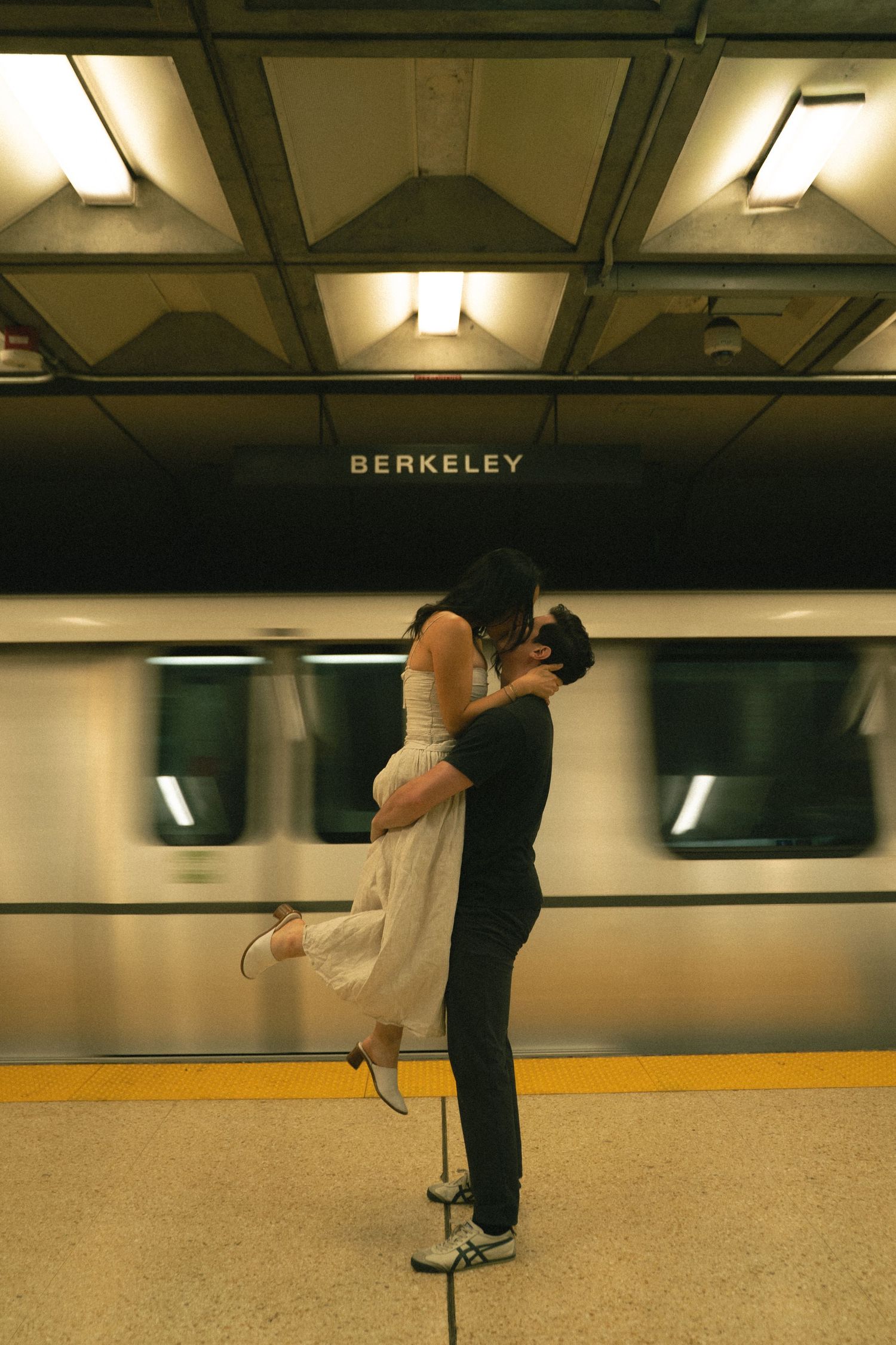 Couple kissing at Berkeley BART station with motion-blurred train passing behind them.