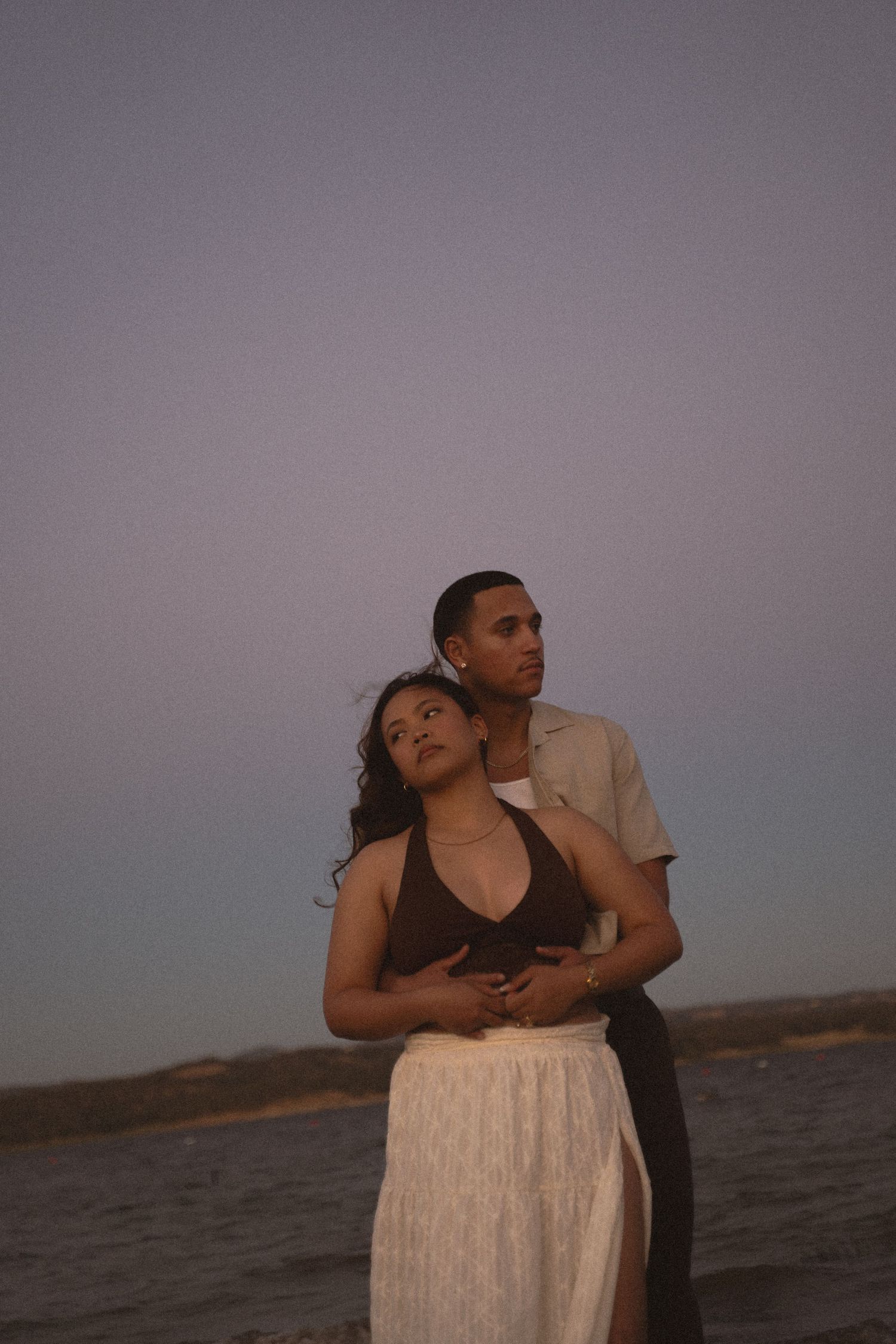 Couple embracing at dusk on a beach, woman leaning back against man under a soft purple sky.