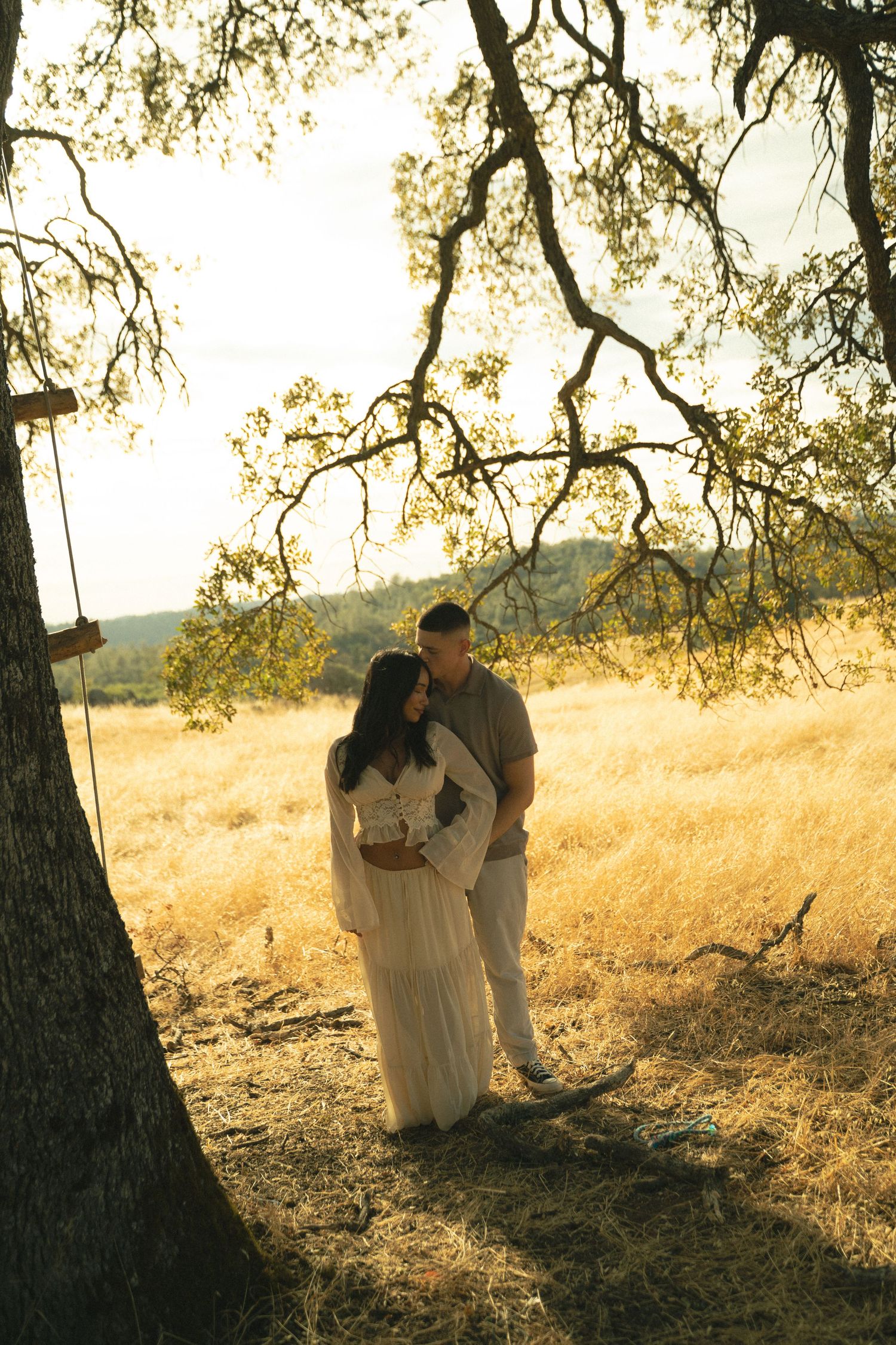 Couple embracing under oak tree in golden sunlit field, woman in white dress with swing nearby.