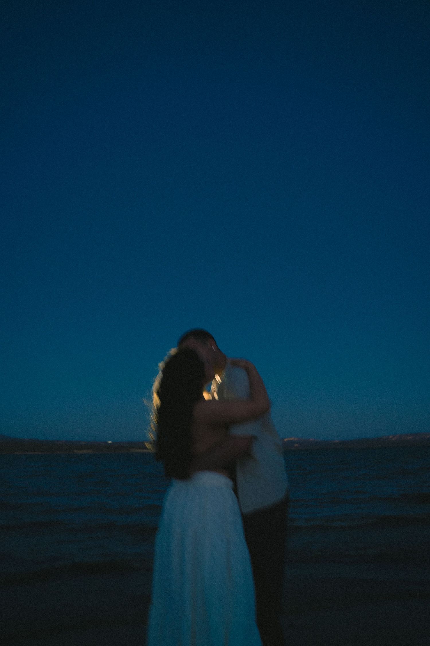 Couple embracing romantically on a beach at twilight with deep blue sky and calm ocean waters.