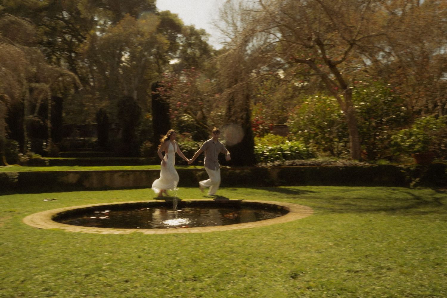 Couple holding hands and jumping near a circular garden fountain surrounded by manicured green lawn.