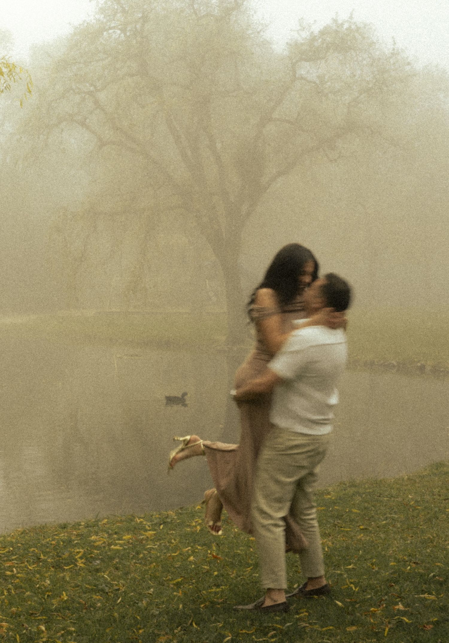 Couple embracing romantically by a misty lake with a large tree in the foggy background.