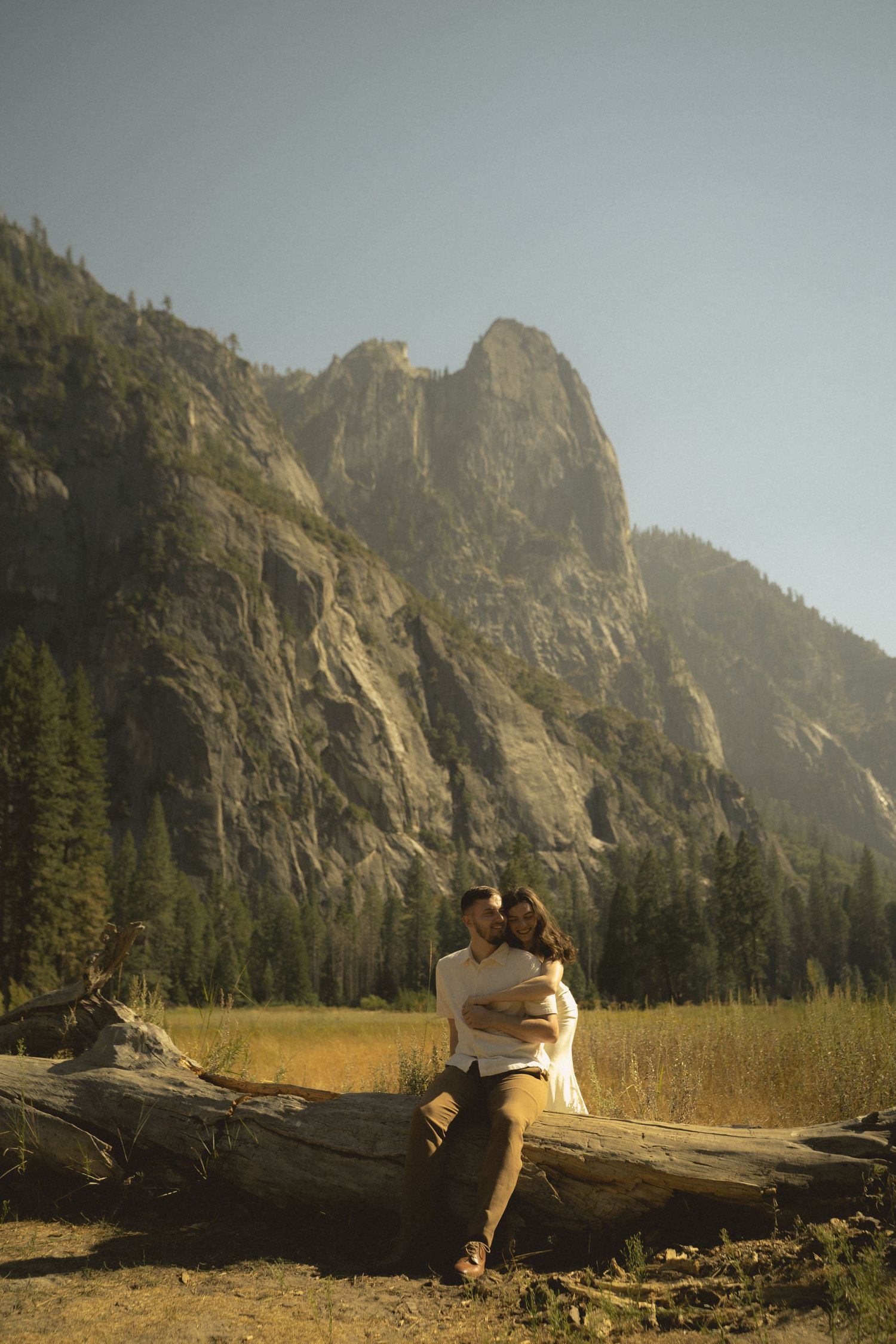 Couple embracing on a fallen log with dramatic granite mountains and pine trees in Yosemite National Park.