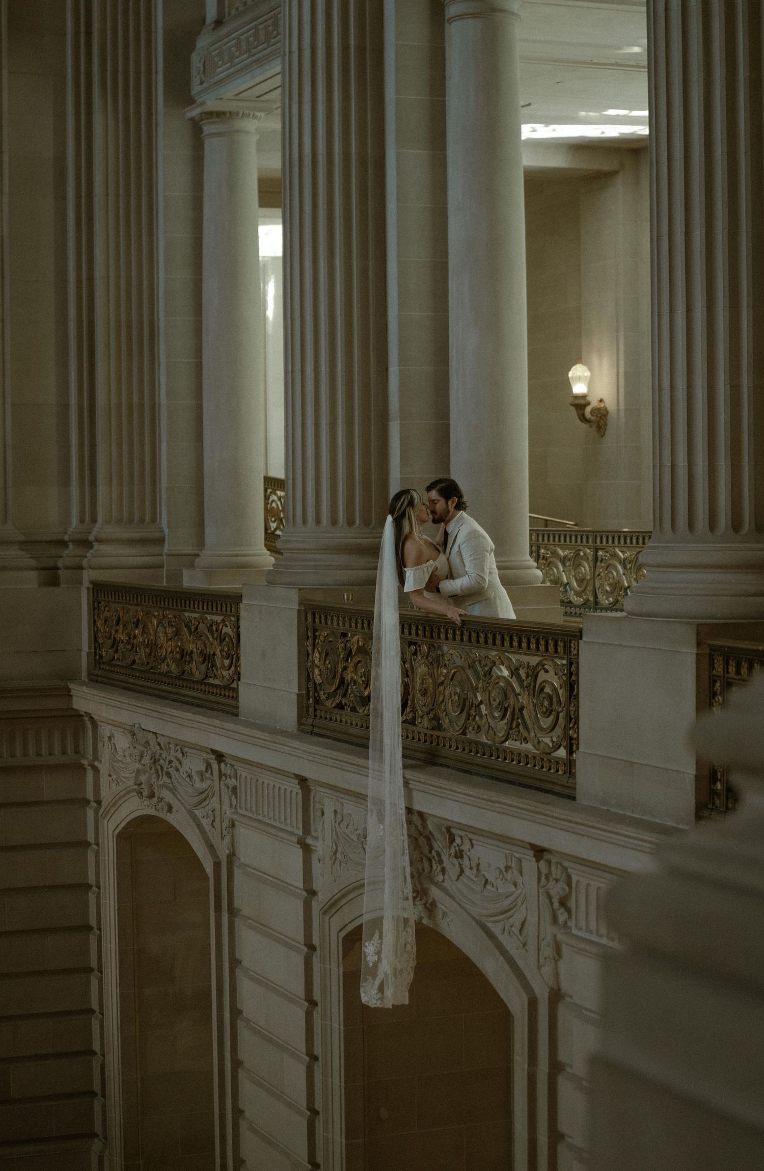 A bride and groom share a kiss on an ornate balcony with elegant columns in a grand wedding venue.