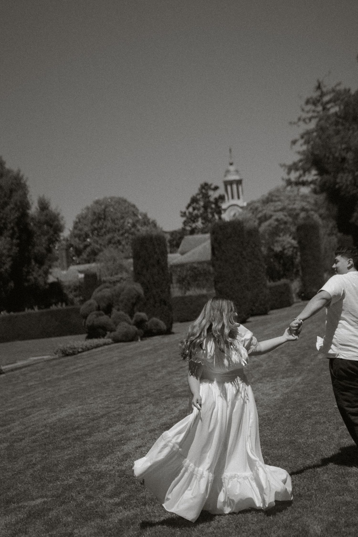 Black and white photo of a bride in a flowing dress holding hands with her groom in a garden.