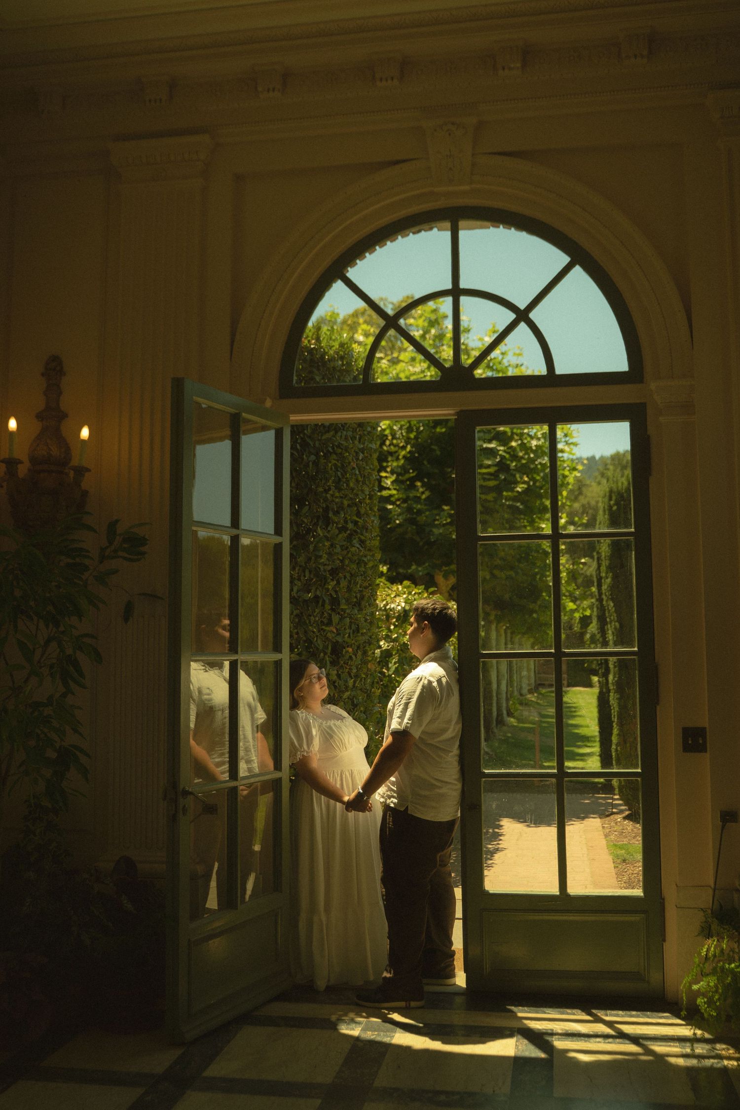 Bride and groom holding hands in grand doorway with arched window overlooking sunlit garden path.