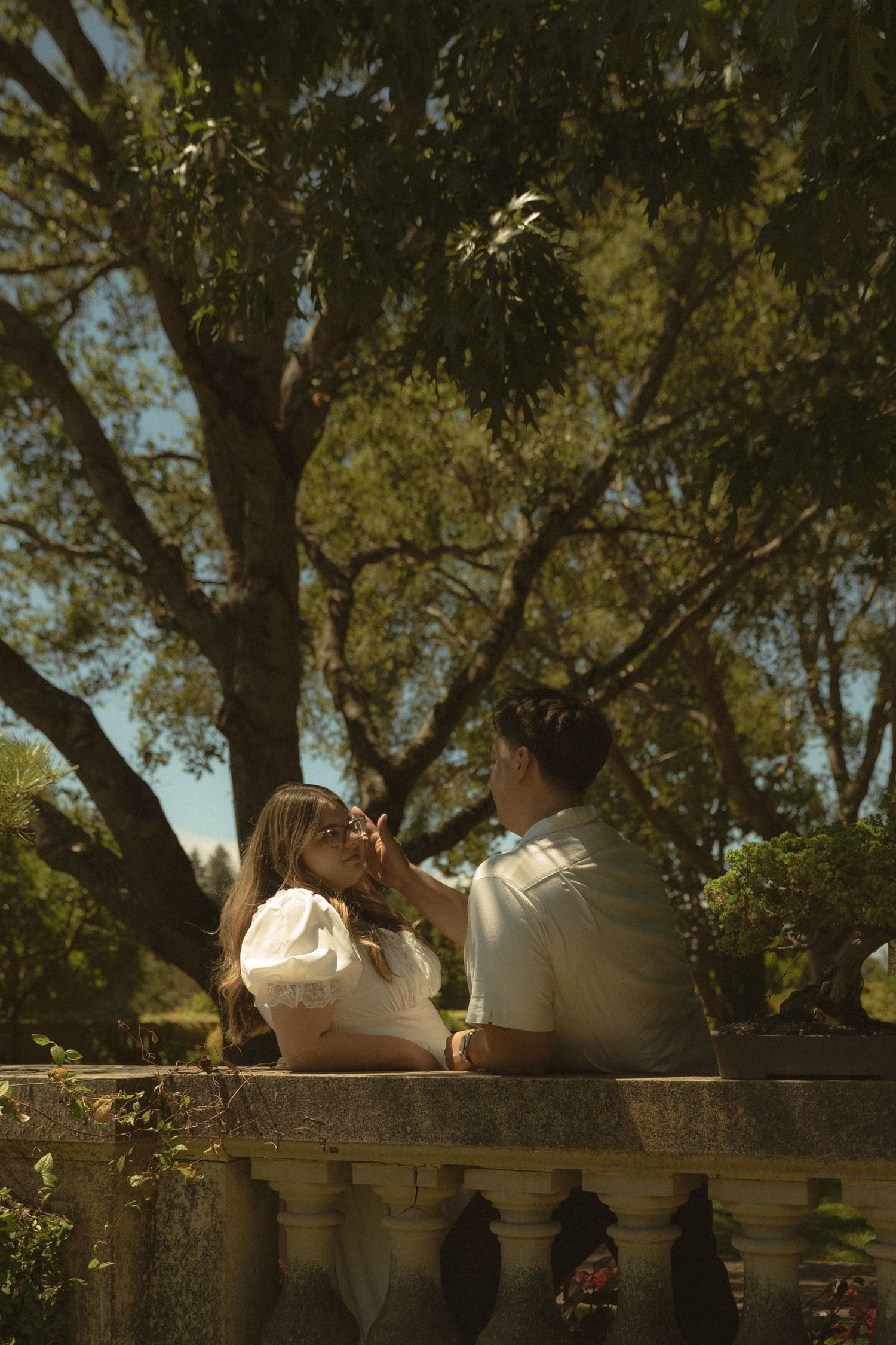 Romantic couple sitting on stone balustrade sharing tender moment beneath large shady garden trees.