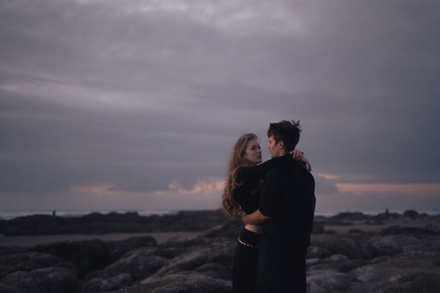 A couple holds each other close on a rugged coastline with a dramatic cloudy sunset sky behind them.