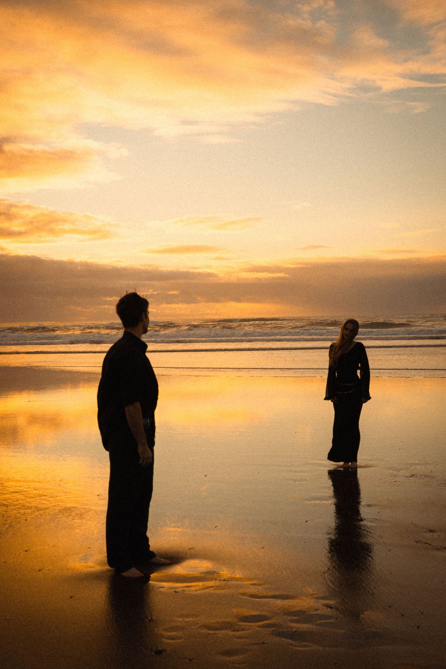 Two people in black stand apart on a reflective beach at sunset with a dramatic golden sky.